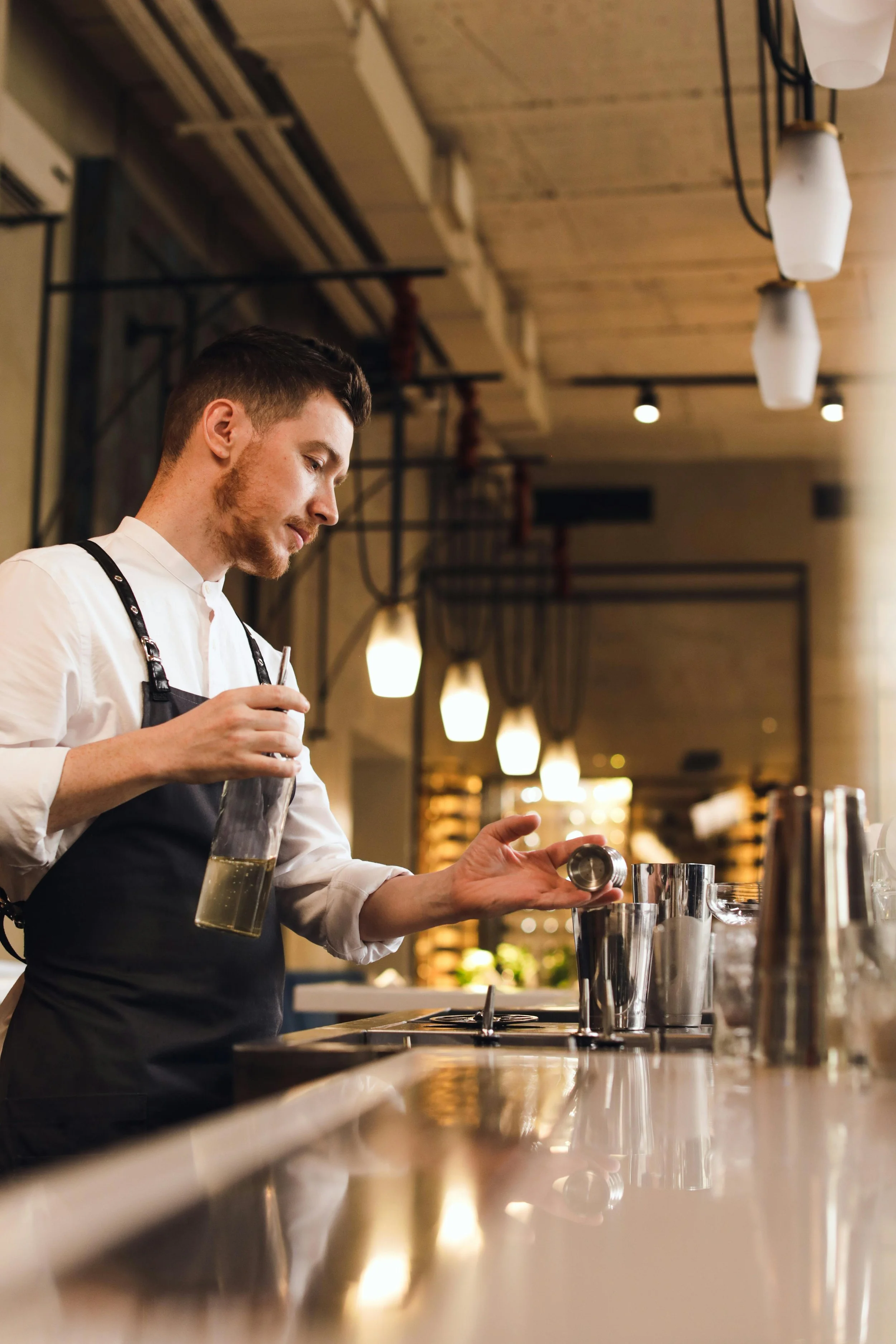 Bartender preparing a drink with a jigger and bottle behind a bar counter.