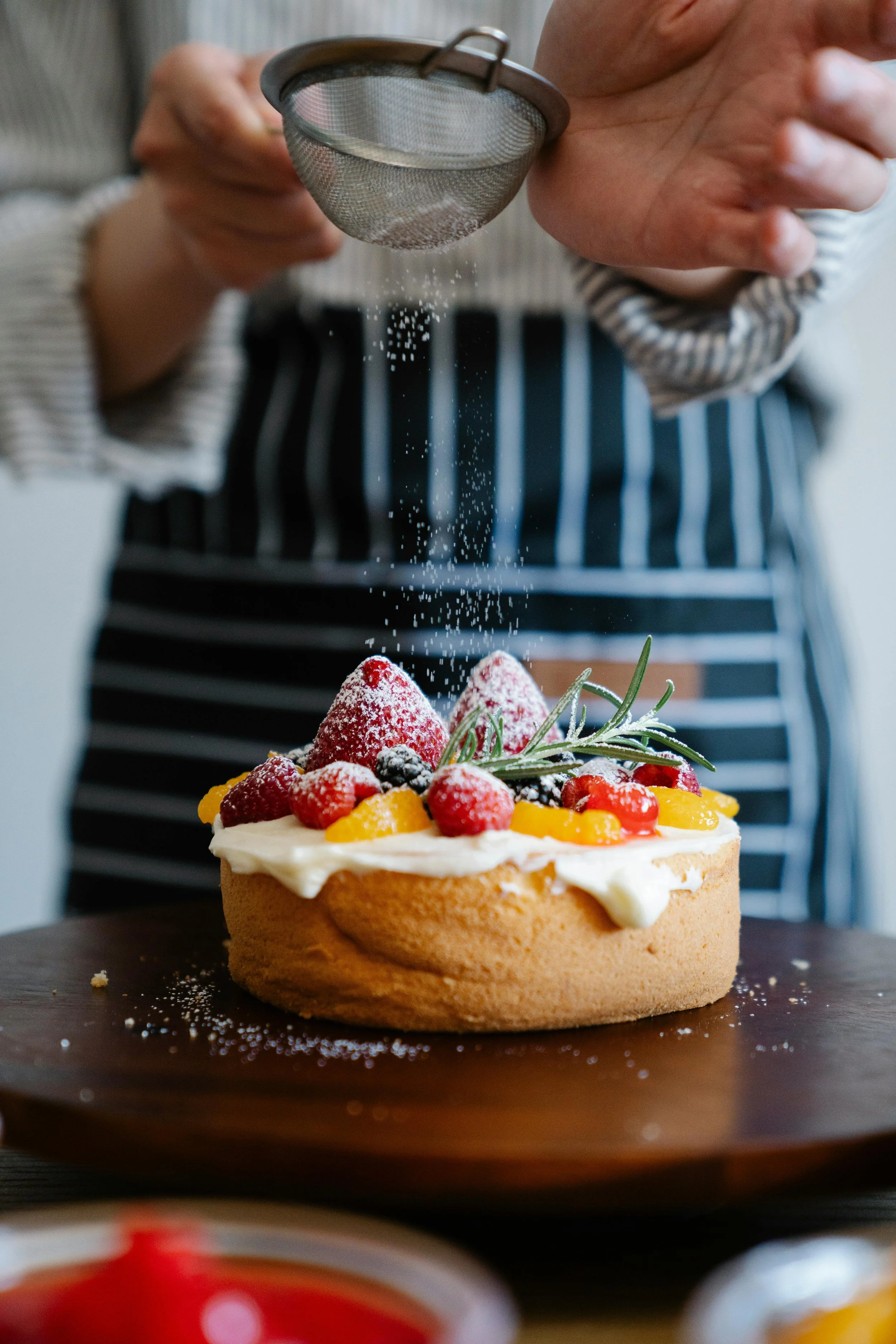 Person sprinkling powdered sugar on cake topped with fruit