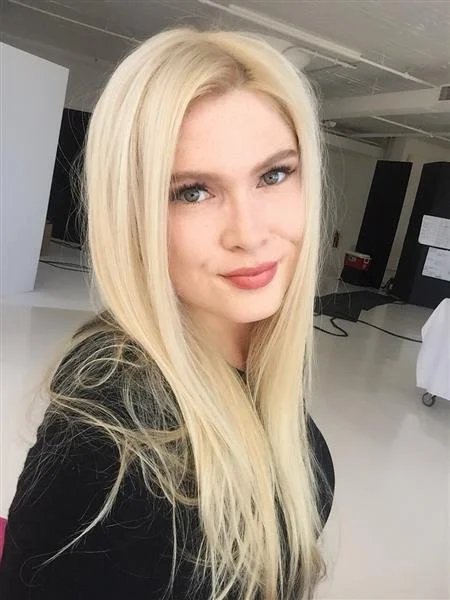 Woman with long blonde hair smiling in a brightly lit room with a black top and natural makeup.