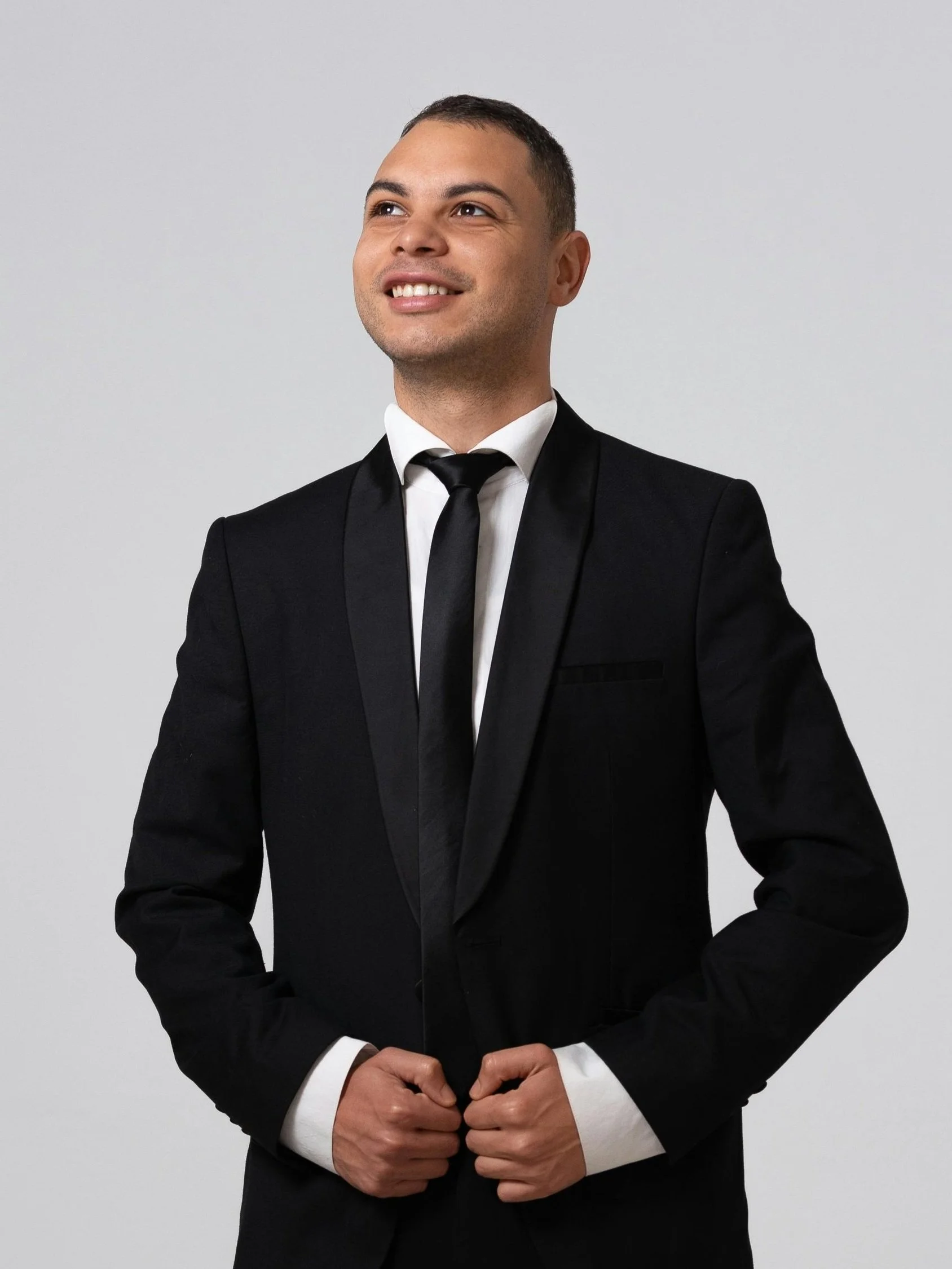 Man in a black suit and tie smiling, looking upwards, against a plain background.