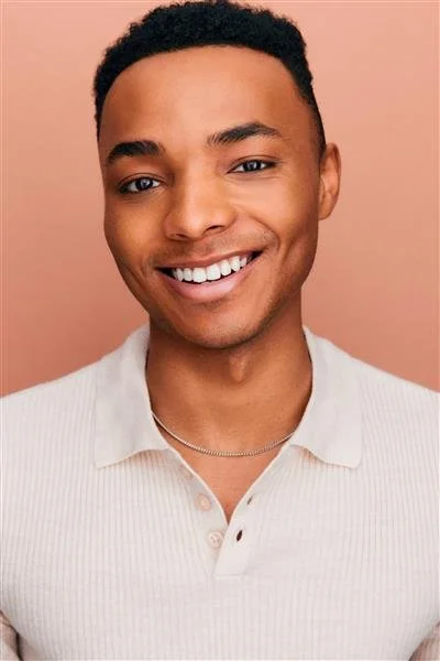 Smiling person wearing a light-colored collared shirt against a peach background.