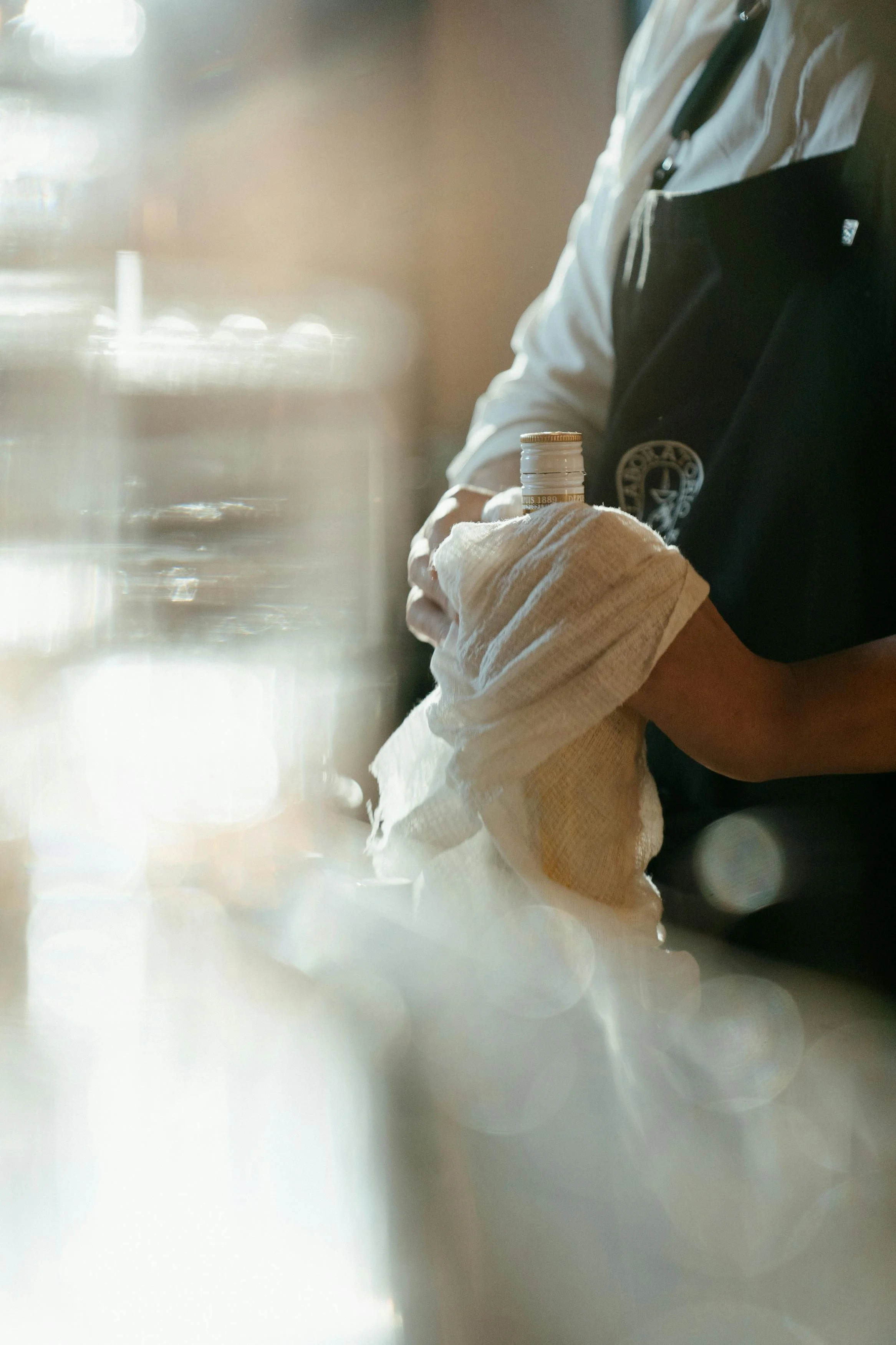 Person polishing a bottle with a cloth, wearing a dark apron.