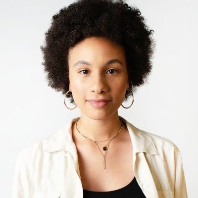 Person with curly hair and hoop earrings, wearing a cream-colored shirt and necklaces, against a plain background.