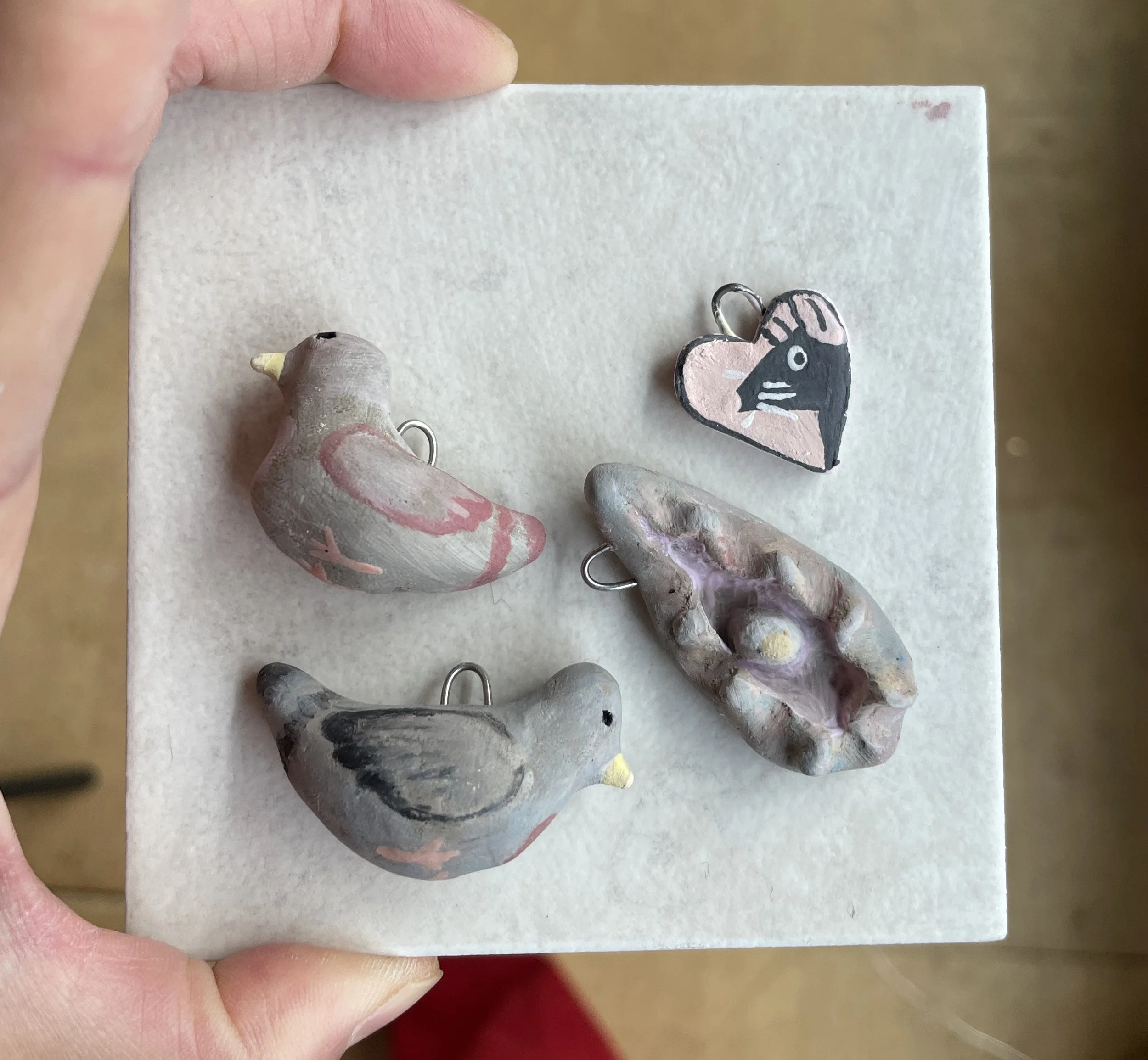 Hand holding a white display board with four painted clay bird charms and one painted heart-shaped charm.