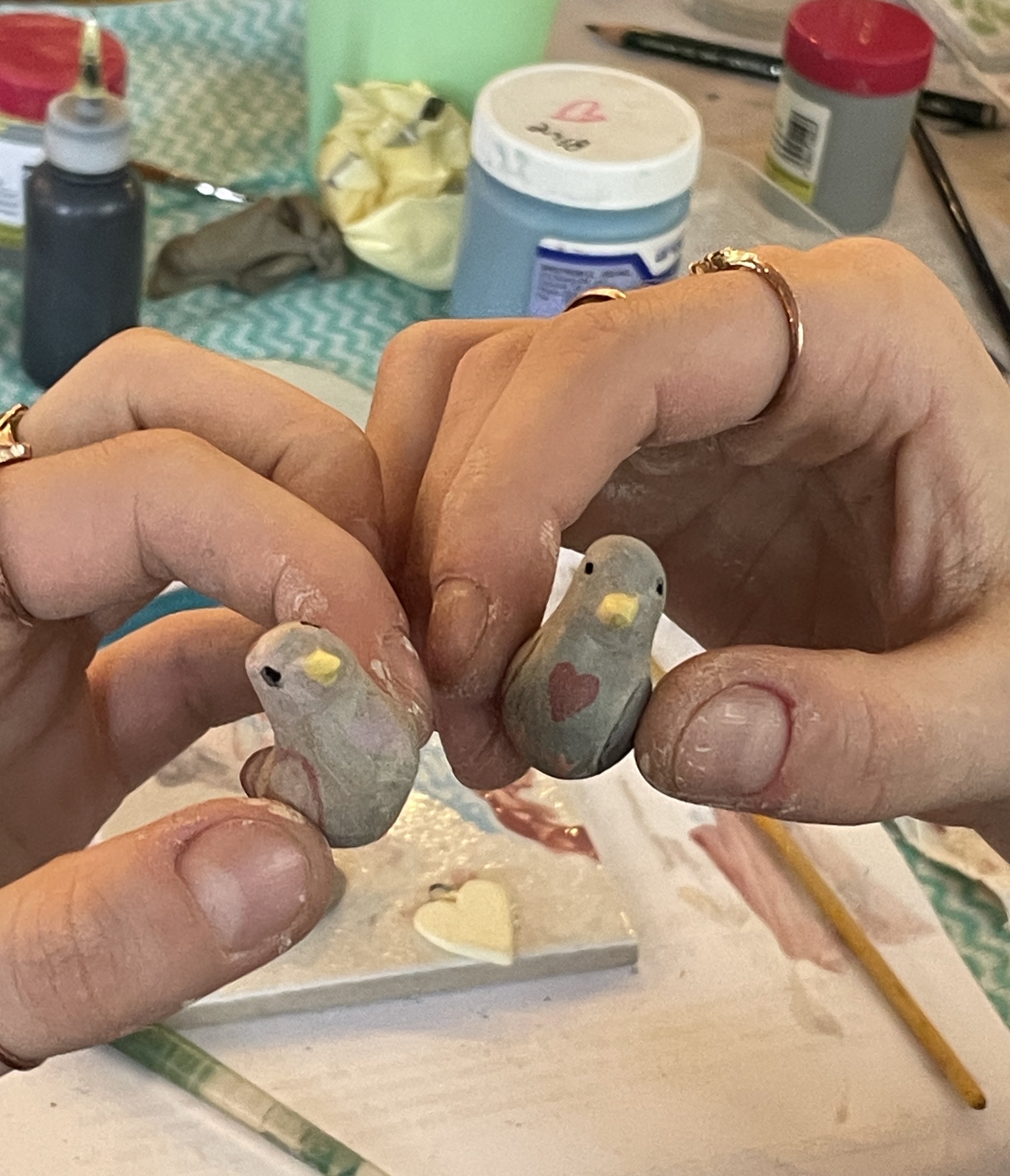 Person holding two small painted bird figurines with hearts on their chests, surrounded by art supplies on a table.
