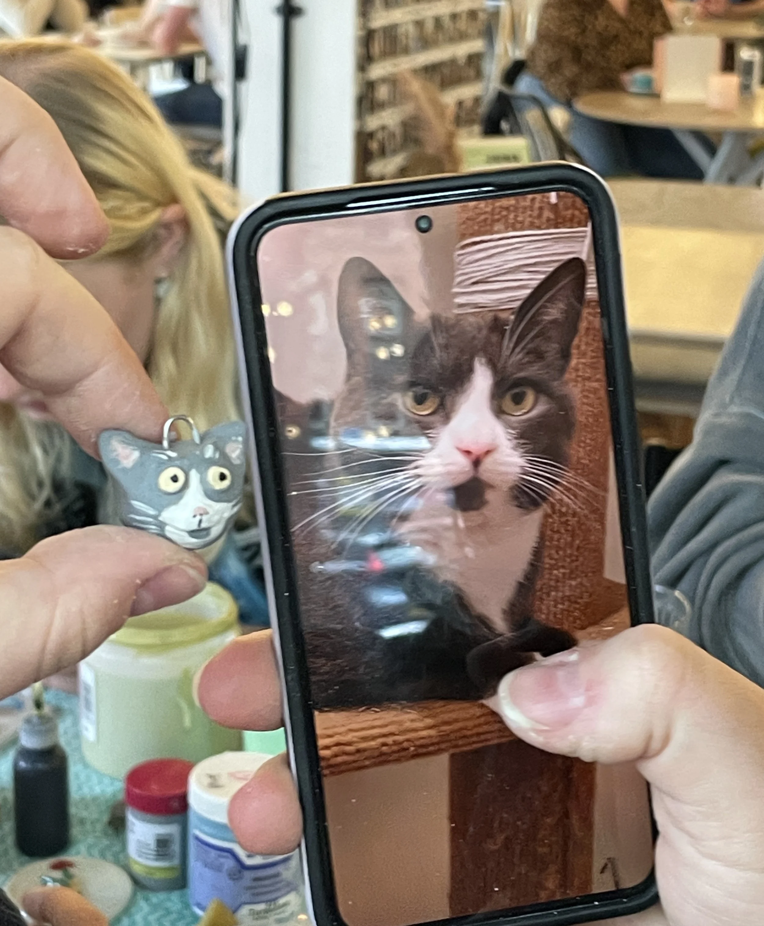 A person holds up a smartphone displaying a photo of a black and white cat with yellow eyes, sitting on a wooden surface. In the background, a hand holds a gray cat-shaped charm with yellow eyes.