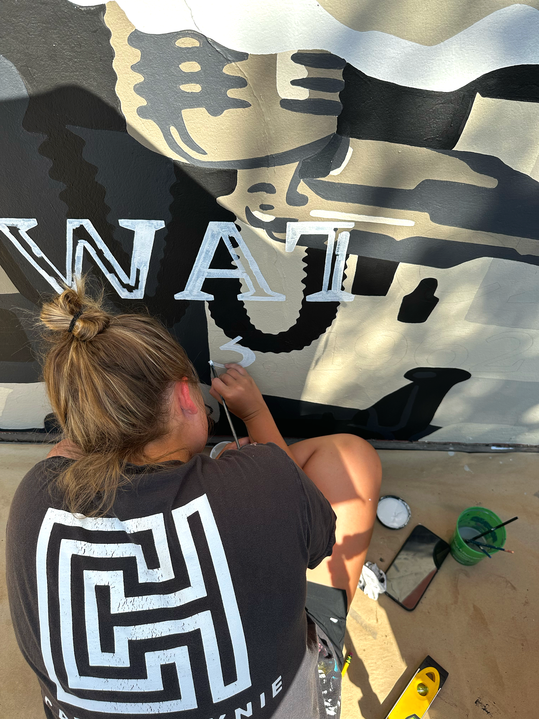 A person with a bun hairstyle painting a mural on a wall, showing abstract shapes in black, white, and beige colors.