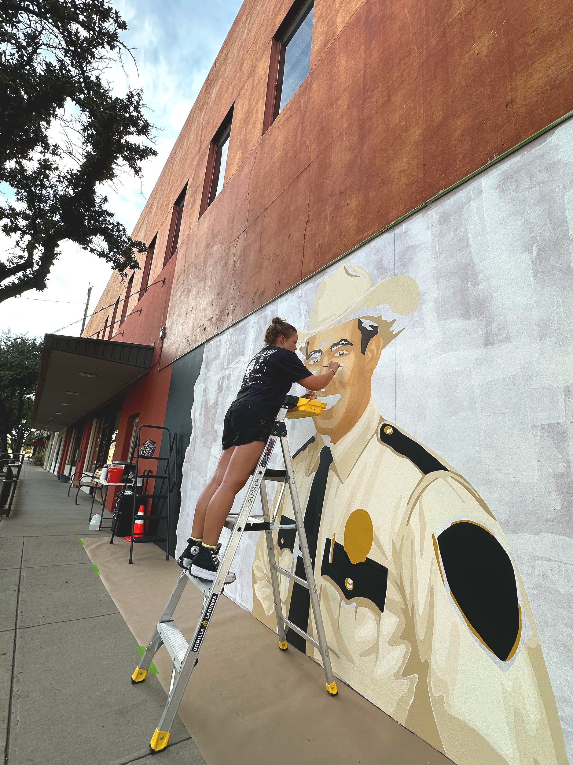 A person standing on a ladder painting a mural of a smiling man in a sheriff or police uniform on a building exterior.