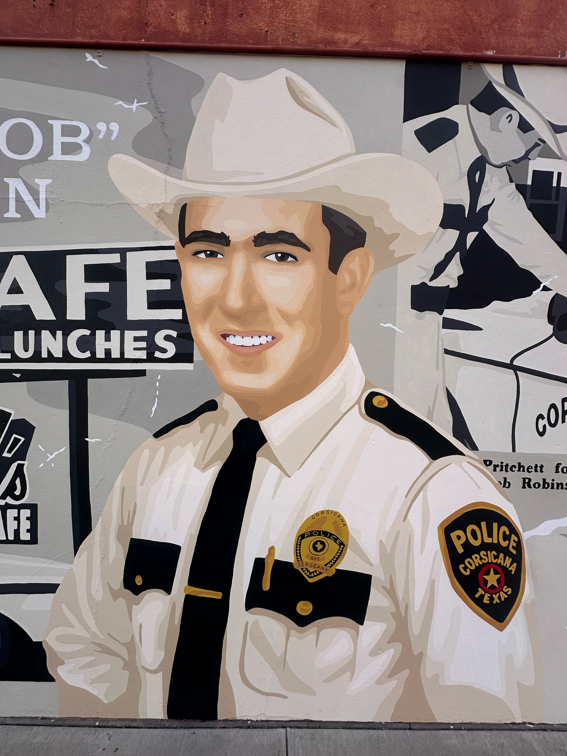A mural of a smiling police officer in a sheriff's uniform, wearing a beige cowboy hat, with police badges and patches visible. The mural includes text and logo elements related to the sheriff's office.