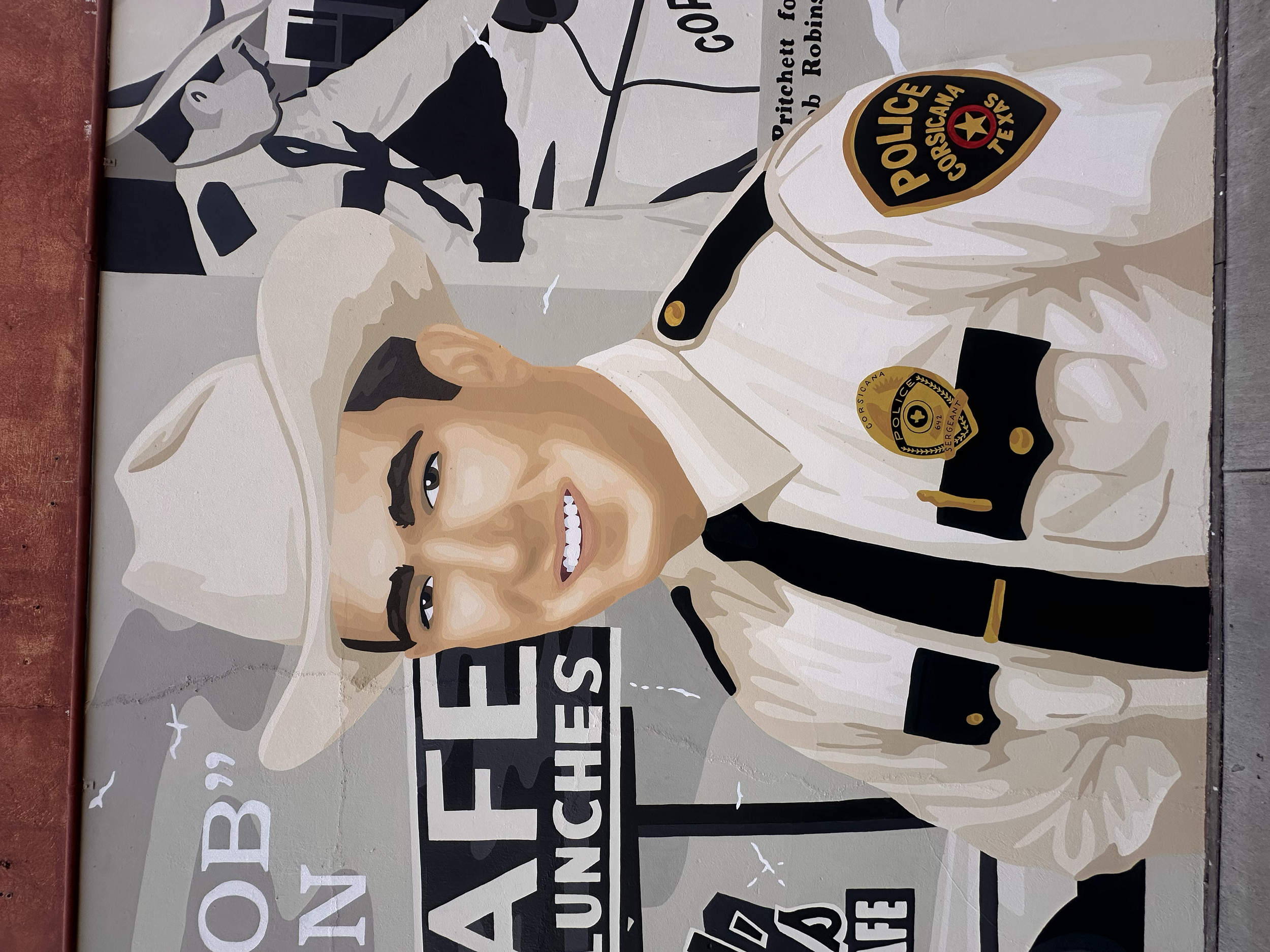 A mural of a smiling police officer in a sheriff's uniform, wearing a beige cowboy hat, with police badges and patches visible. The mural includes text and logo elements related to the sheriff's office.