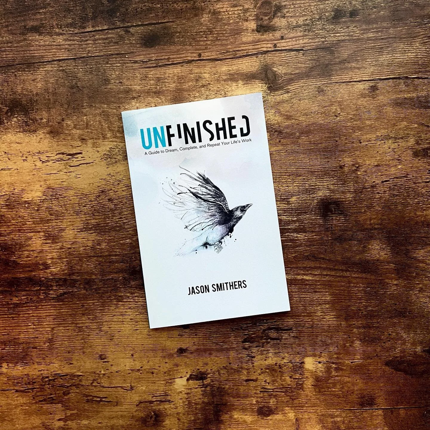 A book titled 'Unfinished' by Jason Smithers, featuring a black and white watercolor illustration of a bird on the cover, placed on a wooden surface.