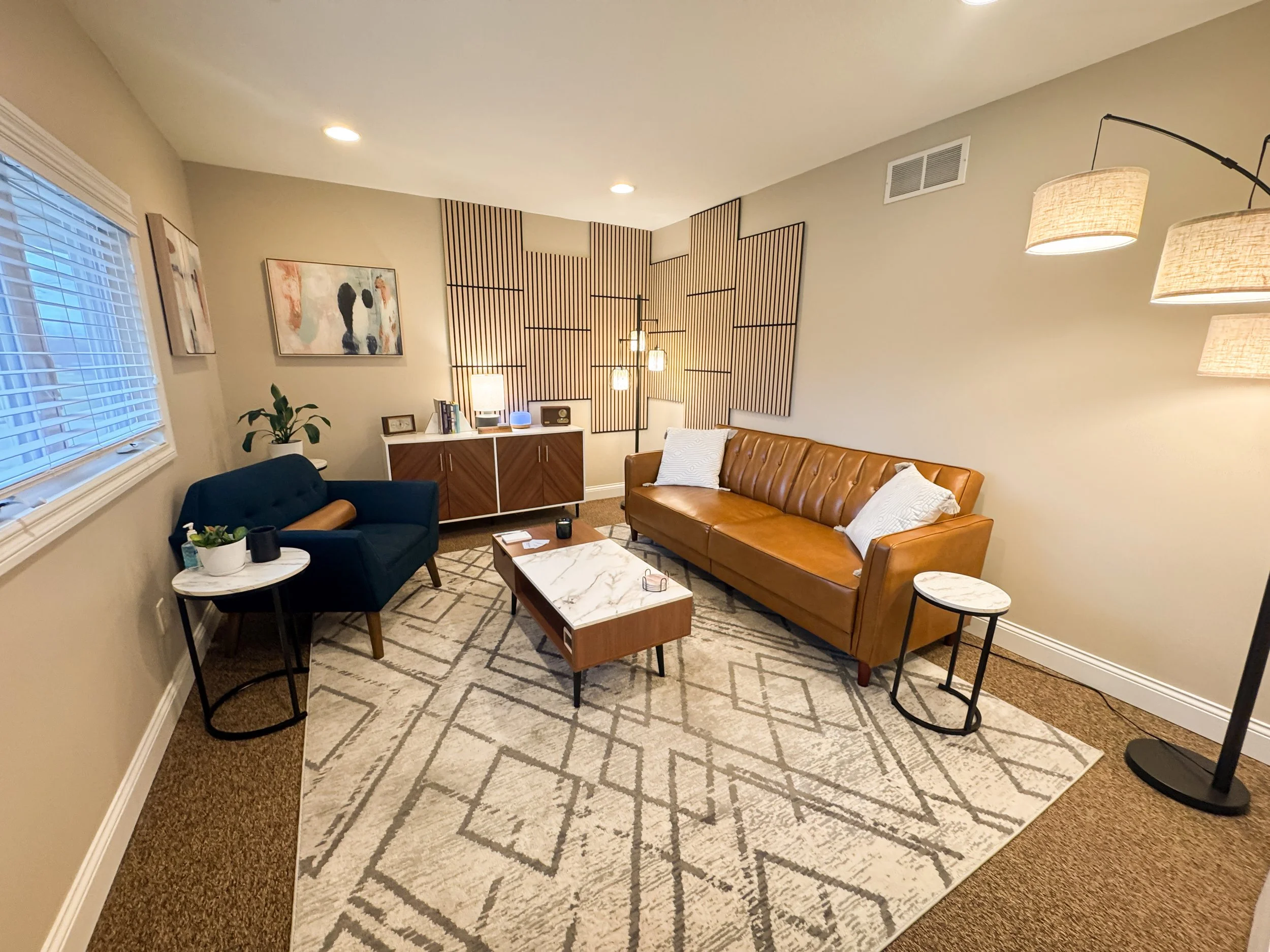 5th Corner Counseling office  with beige walls, a large window with blinds, a brown leather sofa with cushions, a dark blue loveseat, a coffee table with a marble top, a side table, a cabinet with a lamp and decorative items