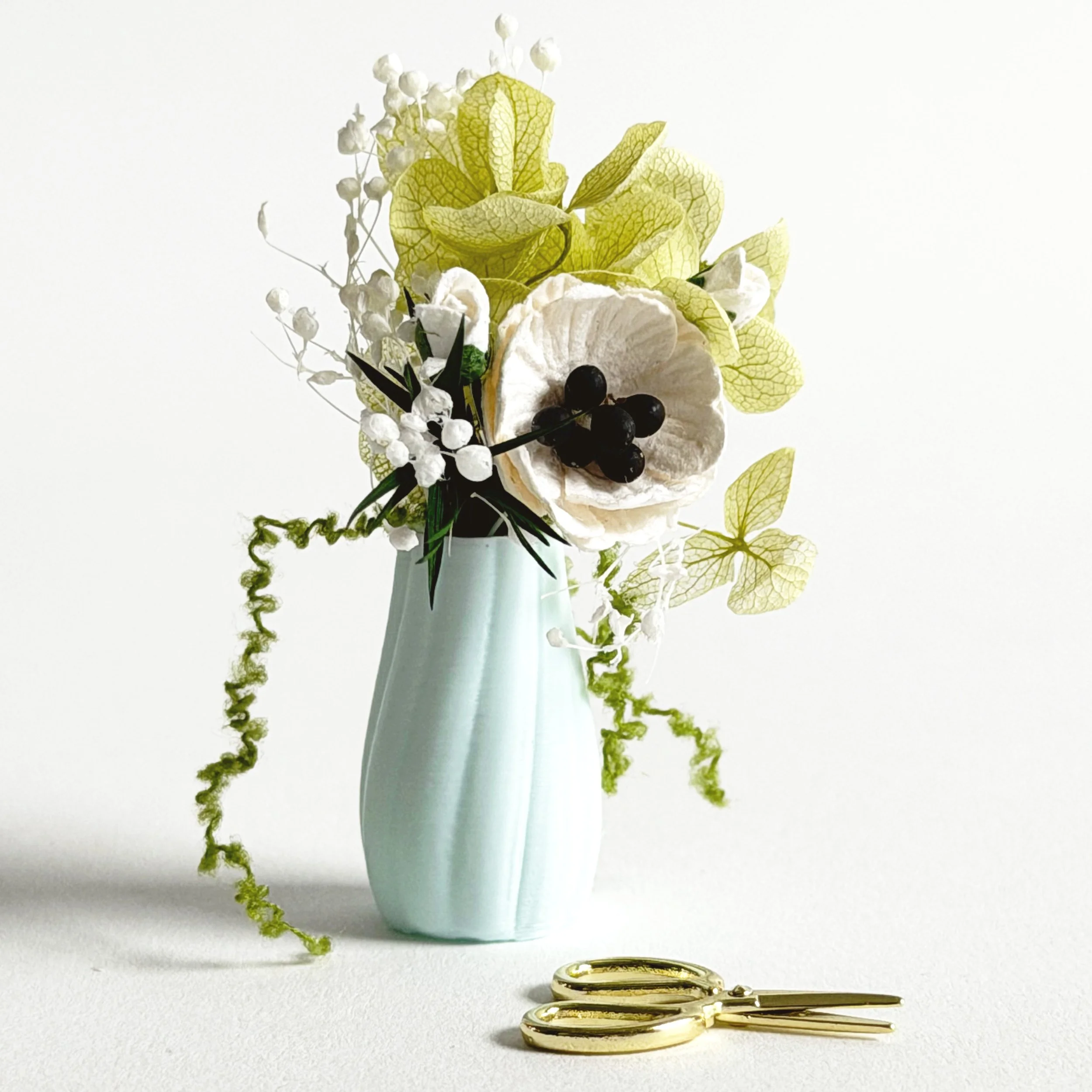 Close-up of green and white miniature floral arrangement with poppies, hydrangea, and trailing greenery in small vase