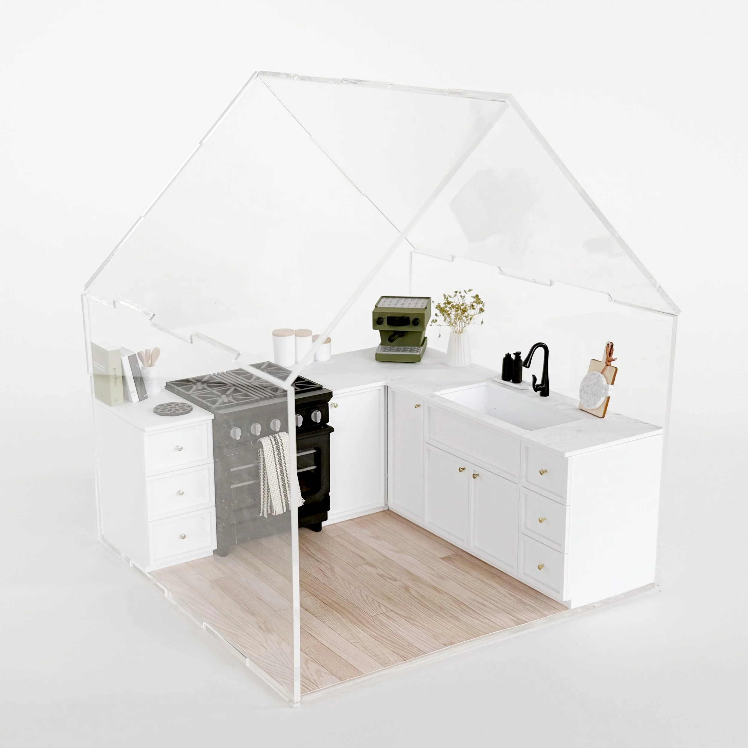 Modern miniature kitchen setup featuring white cabinets and oven, styled inside a clear acrylic dollhouse display.