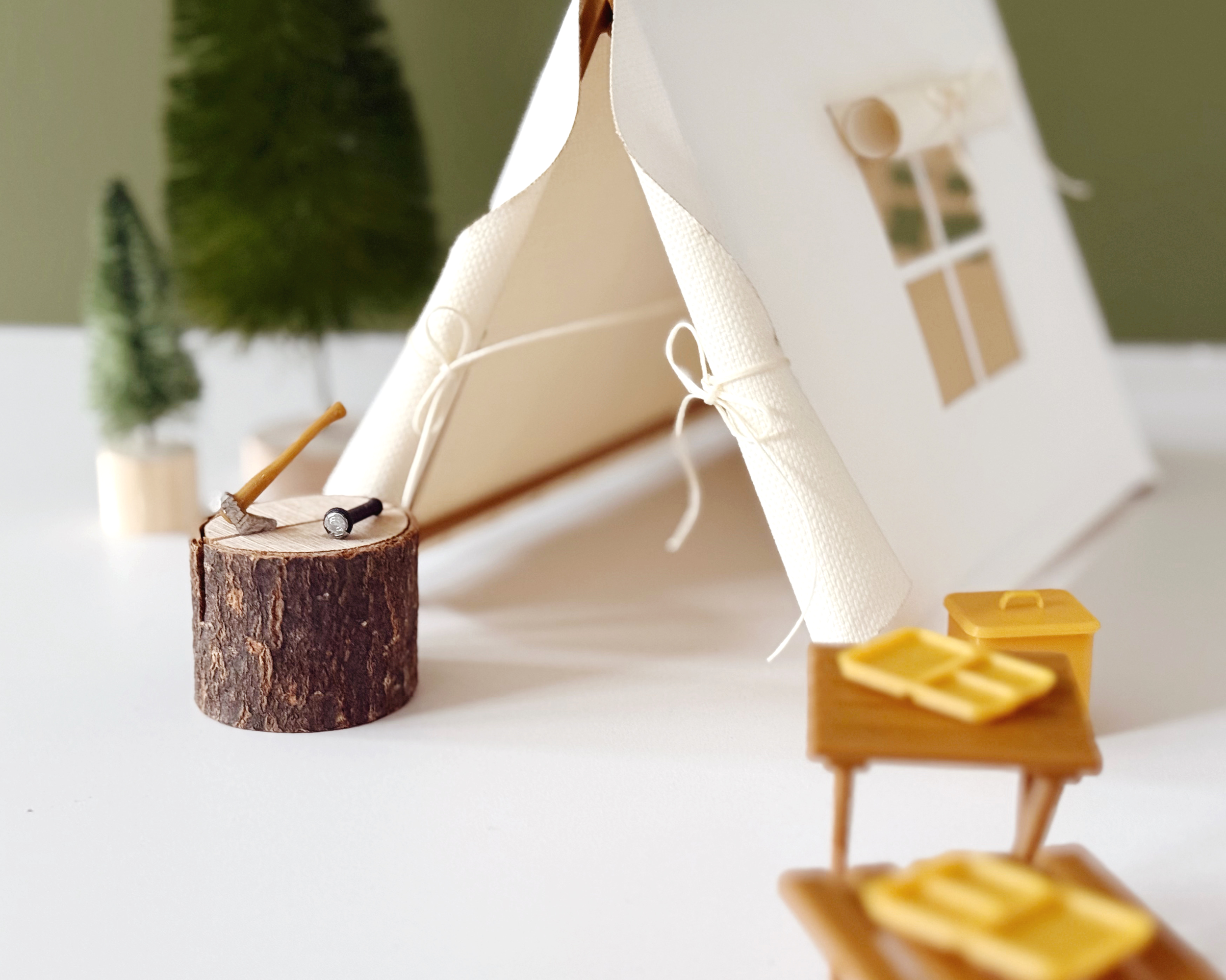 Miniature camping scene with 1:12 scale dollhouse accessories including log, axe, and table next to a tent