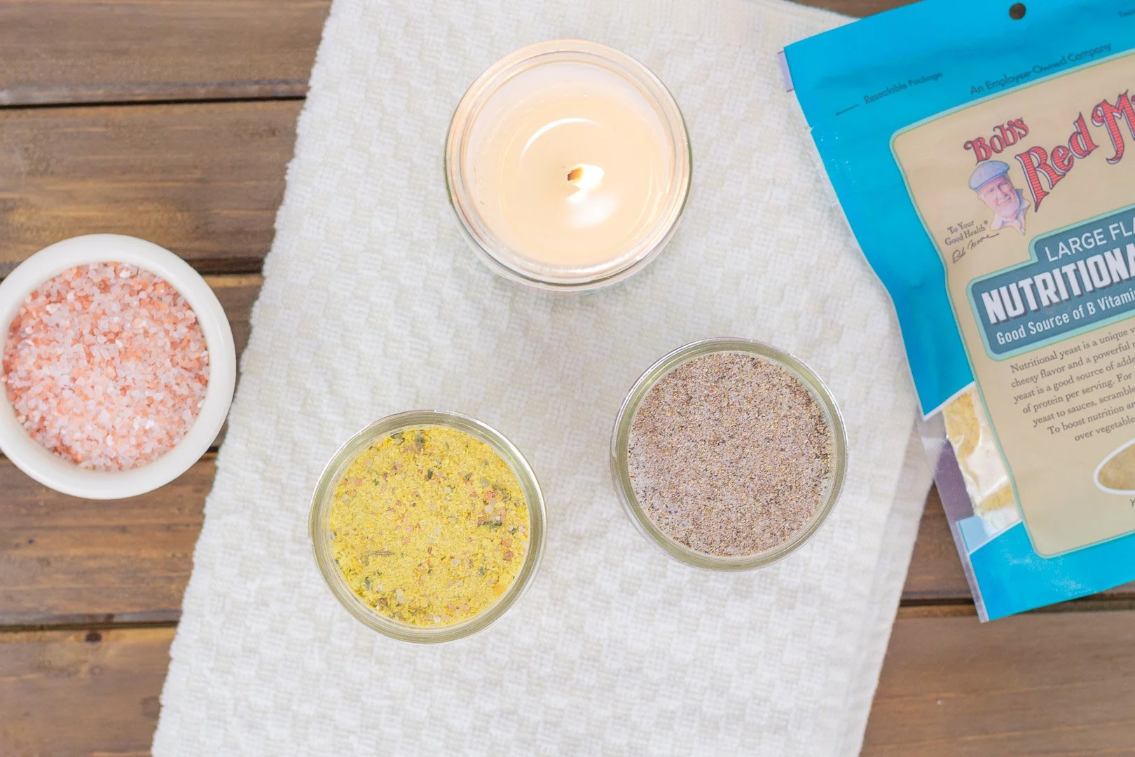 homemade chicken bouillon powder and seasoned salt sitting next to a candle, pink Himalayan salt and Bob's Red Mill Nutritional Yeast