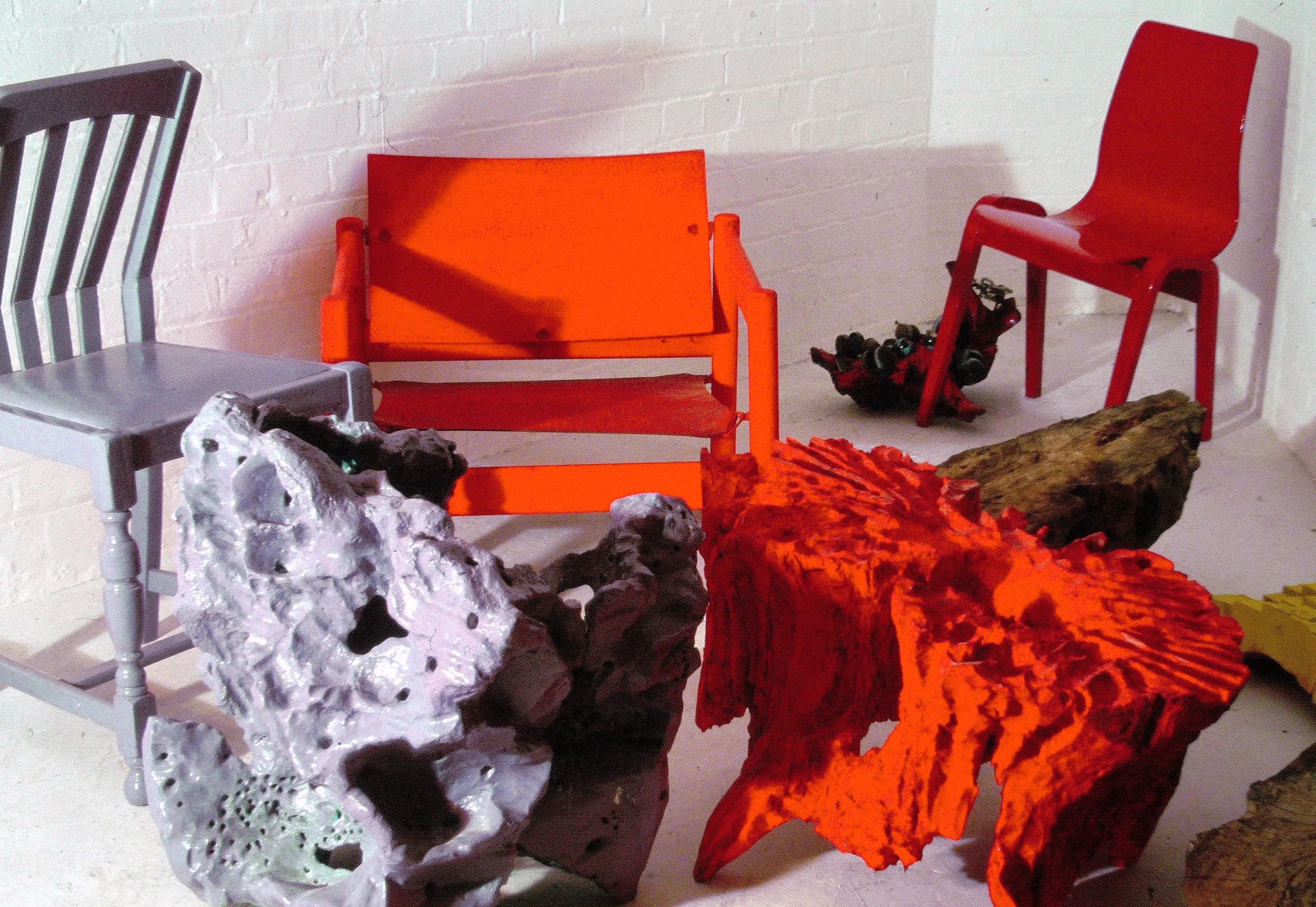 Willcott Synaesthetic artwork, Ba show. Group of colorful chairs, including an orange chair, a red chair, and a gray chair arranged in front of a white brick wall, with various rocks and a piece of wood in the foreground.