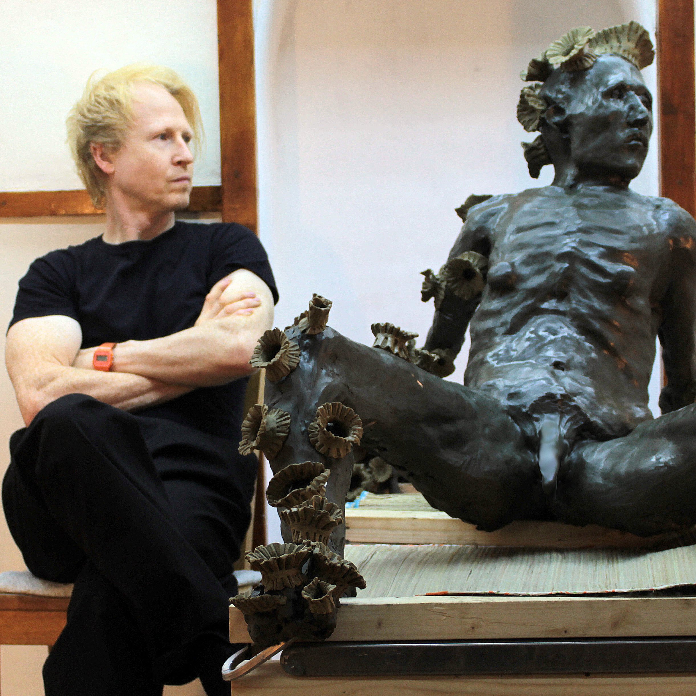 A young man with blond hair, wearing a black shirt and an orange wristwatch, sitting cross-legged on the floor, looking at a bronze bust sculpture of a woman's head with elaborate hair-style and detailed facial features, placed on a wooden table in a