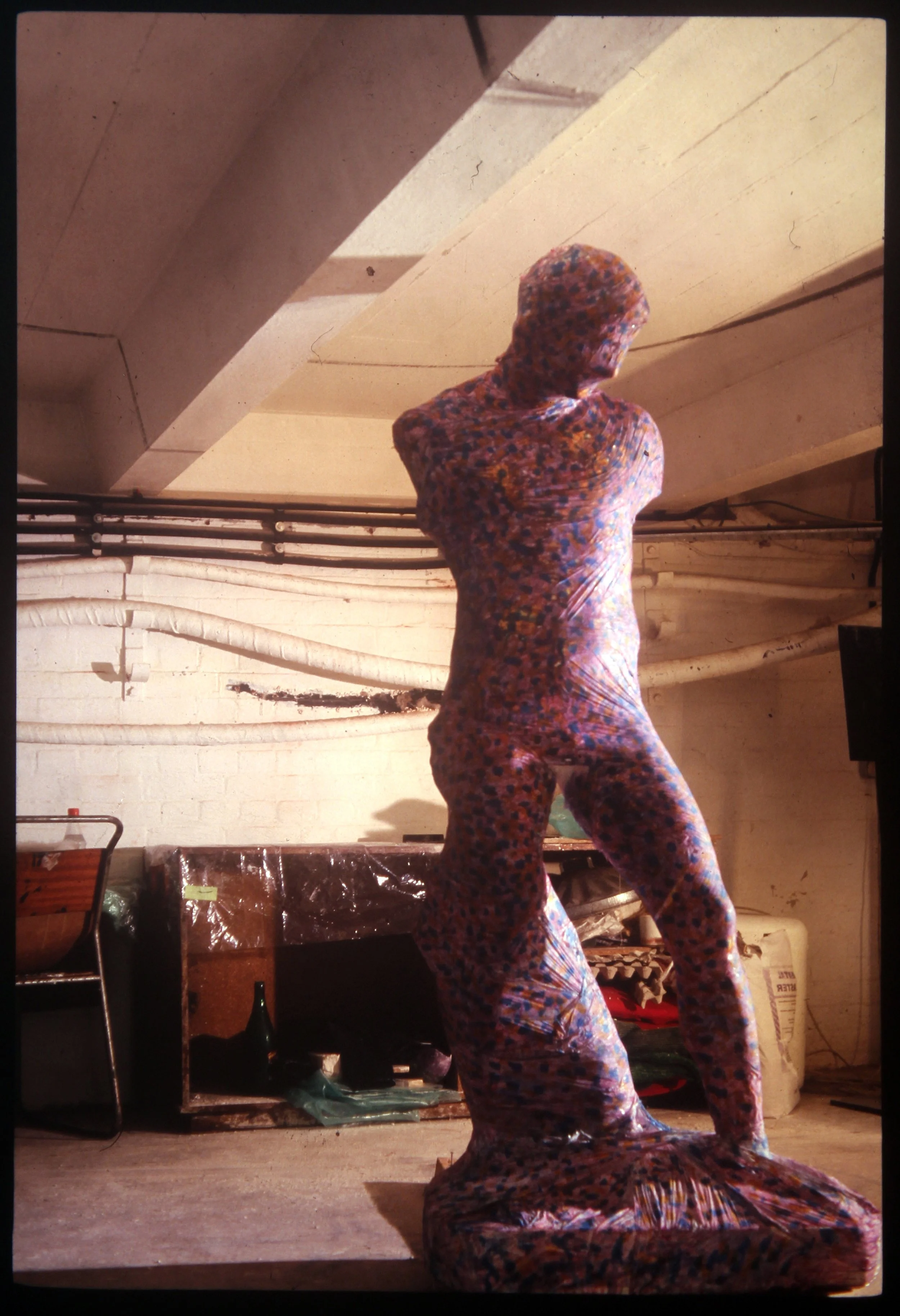 Willcott Synaesthetic artwork, BA degree, person draped in colorful, patterned fabric stands in a room with pipes on the wall and various objects on shelves, facing away from the camera.