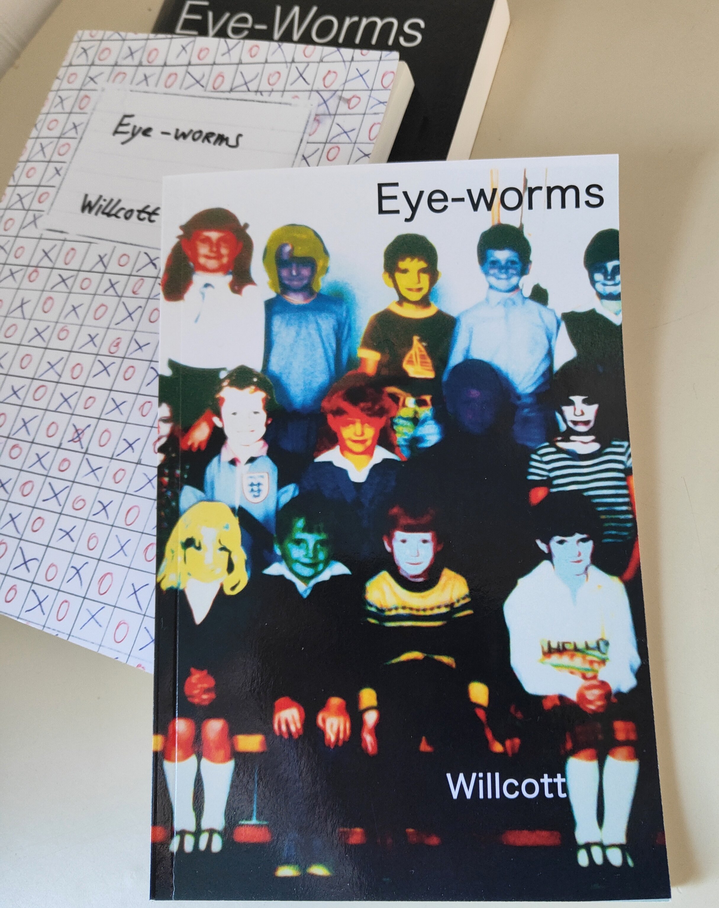 Color-enhanced photograph of a group of children dressed as soccer players, with some in striped uniforms and others in solid colors, standing on a dark background. The text 'Eye-worms' appears at the top, and the name 'Willcott' is at the bottom.