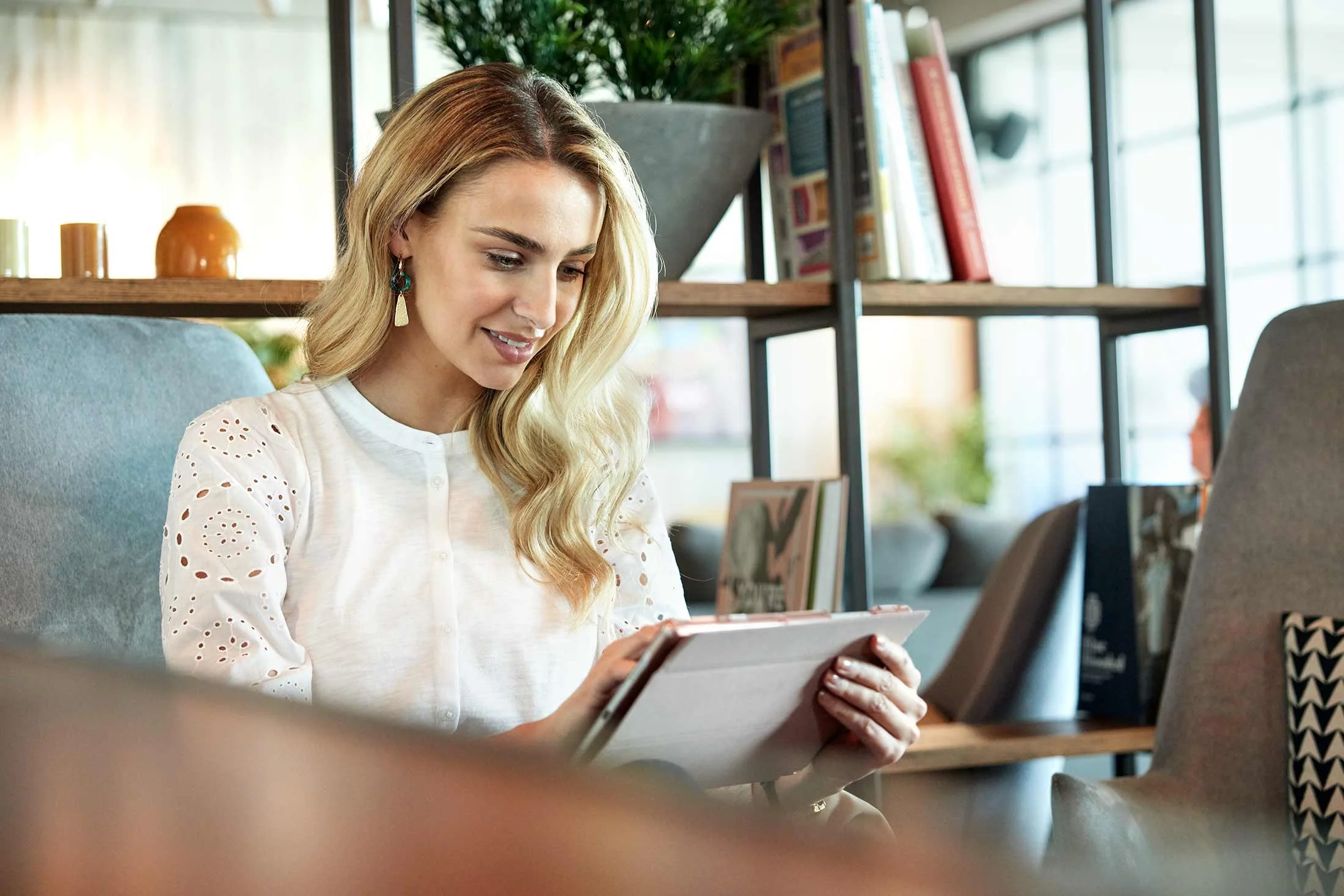 Woman using a tablet in a cozy library setting with books and plants.