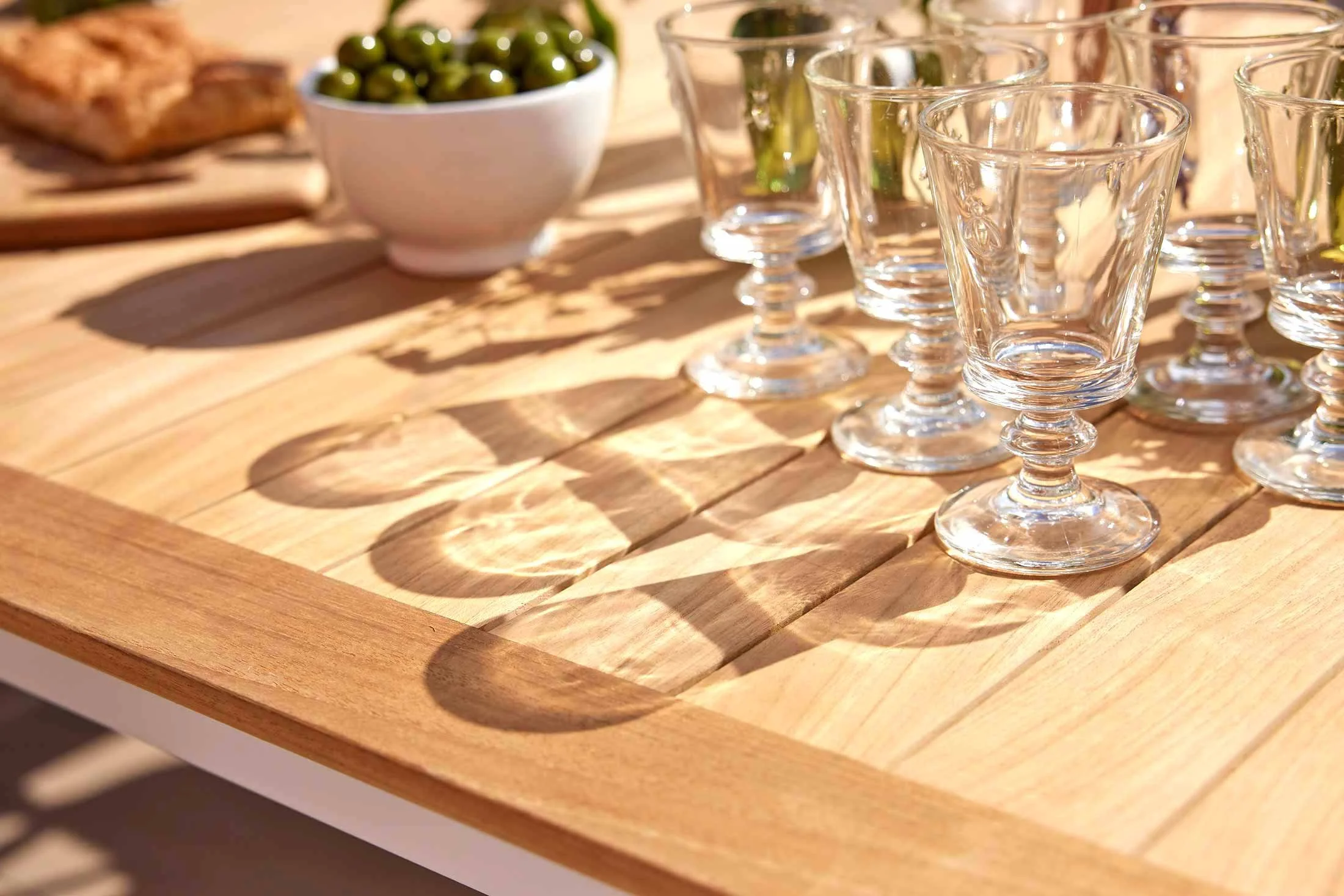 Wooden table with clear glass goblets and a bowl of green olives, casting shadows in sunlight.