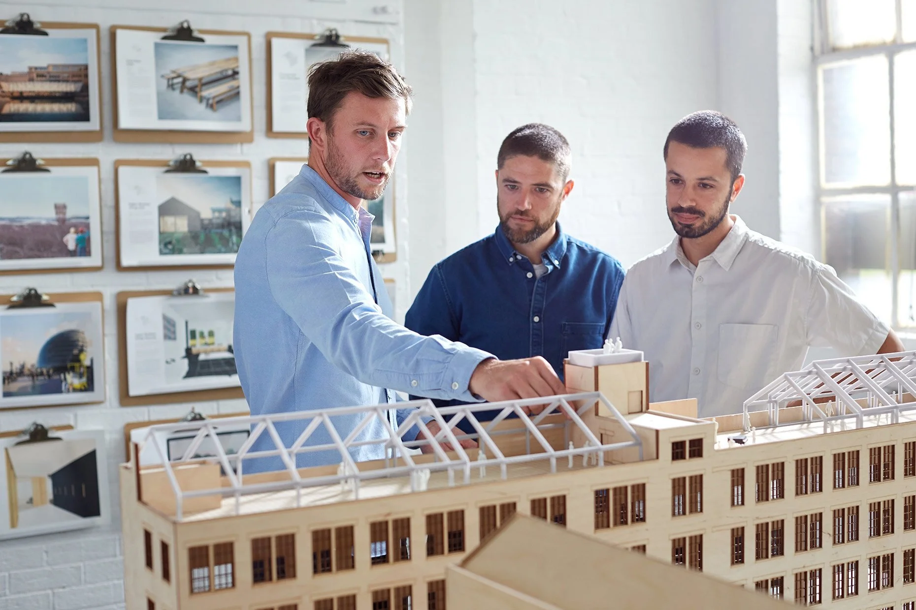 Three architects discussing a building model in an office with architectural designs on the wall.
