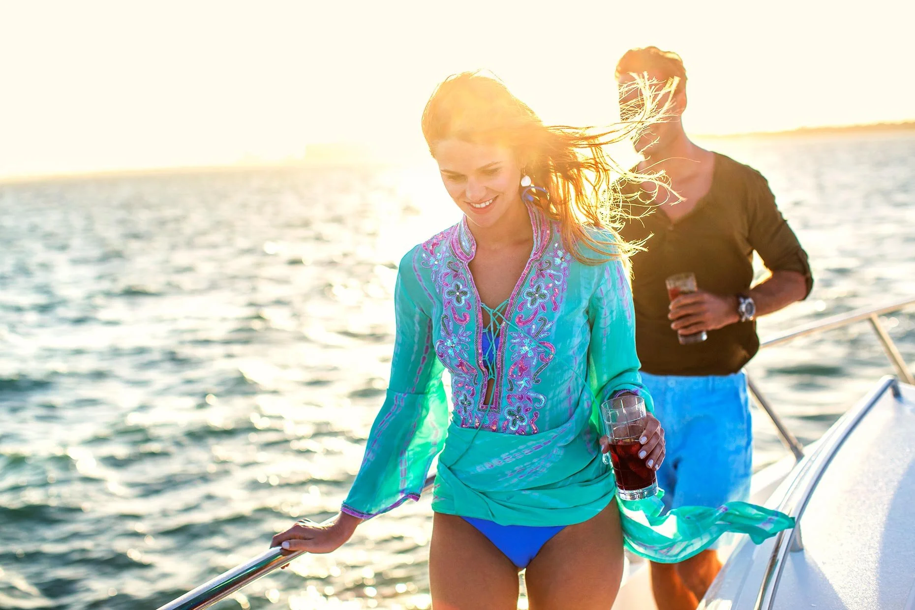 A woman in a colorful tunic and blue bikini bottom holding a drink while walking on a boat, with a man in a black shirt and blue shorts behind her, both enjoying sunny weather on the water.