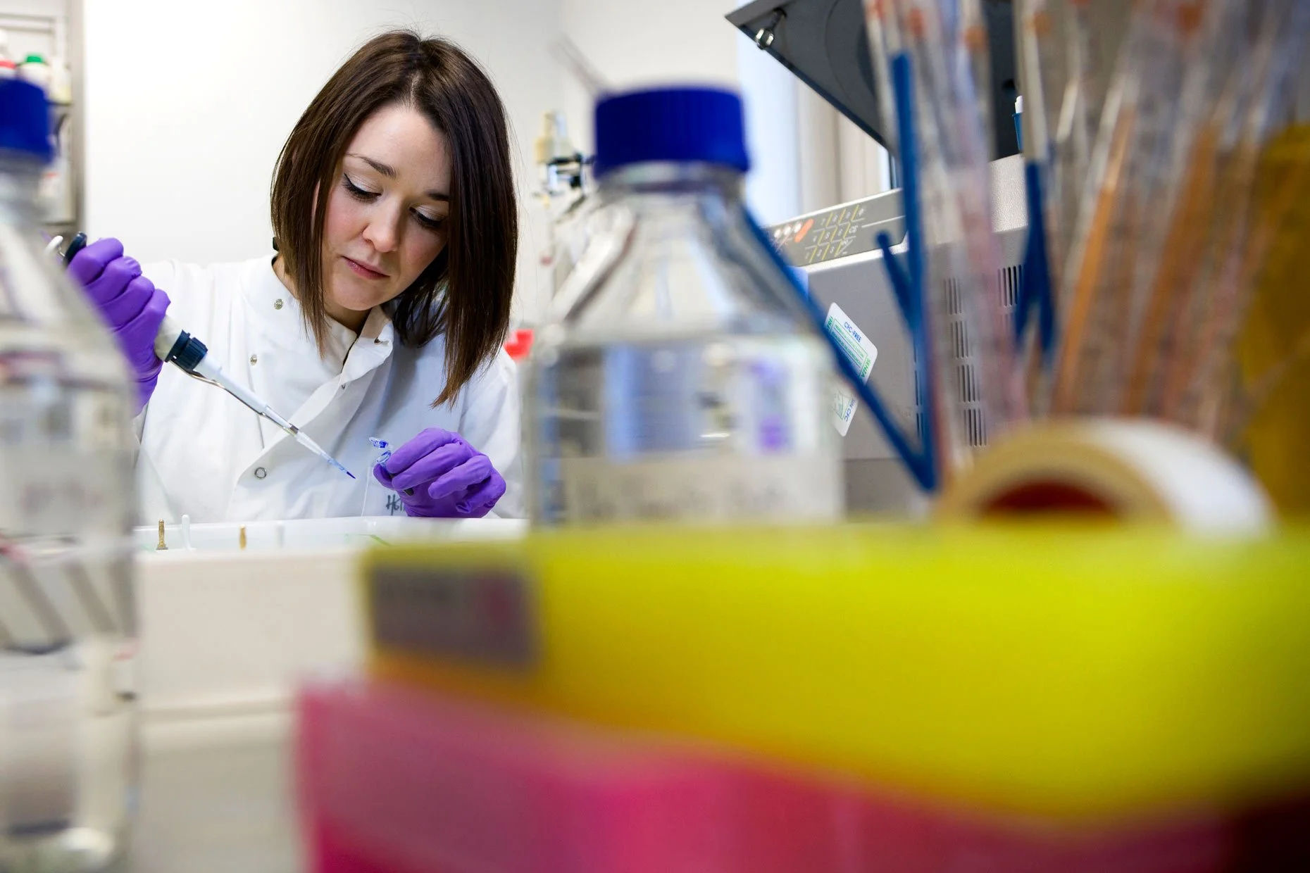 A woman scientist wearing purple gloves and a white lab coat working in a laboratory with various equipment and supplies around her.
