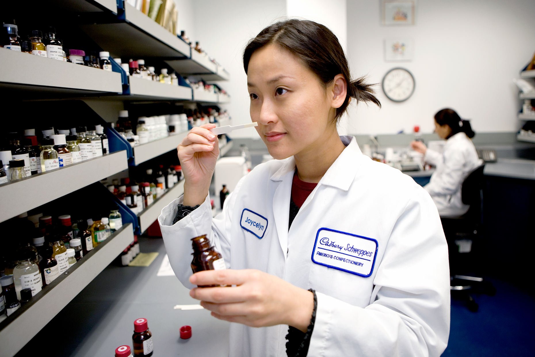A woman in a lab coat holding a small bottle and a dropper in a laboratory, with shelves of bottles on the wall behind her.