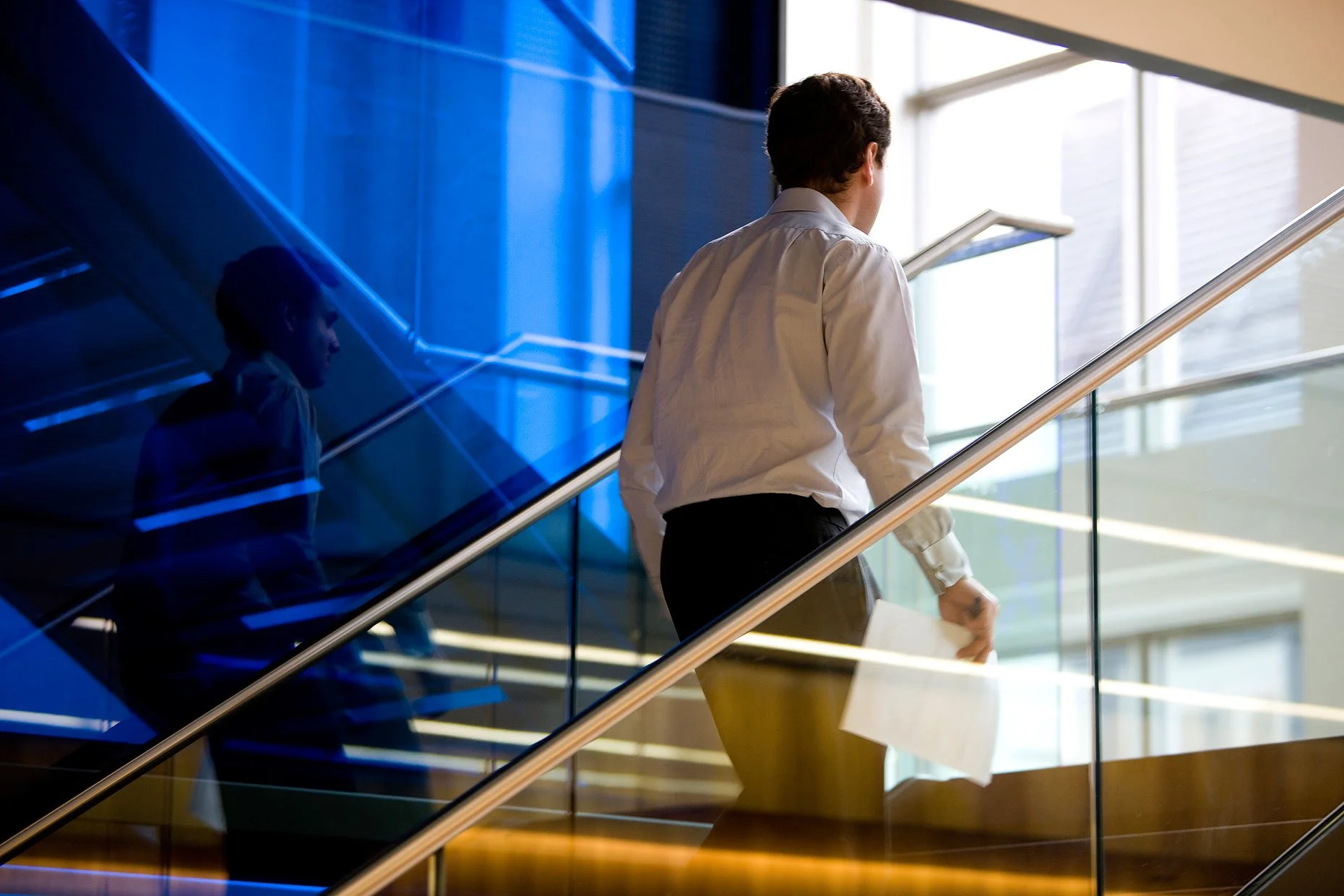 Man in business attire holding paper, ascending a modern glass stairway with blue lighting reflection.