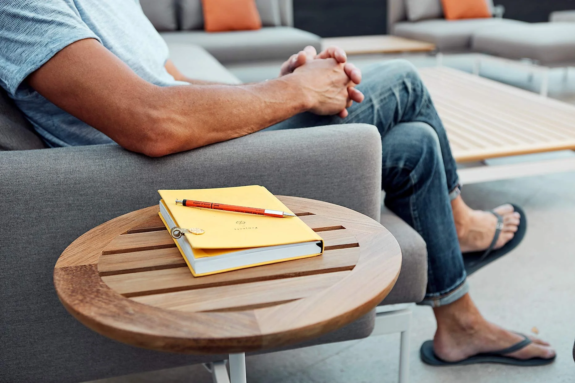 Person seated on a gray sofa with hands clasped, wearing jeans and flip-flops, beside a wooden side table with a closed yellow notebook and an orange pen.