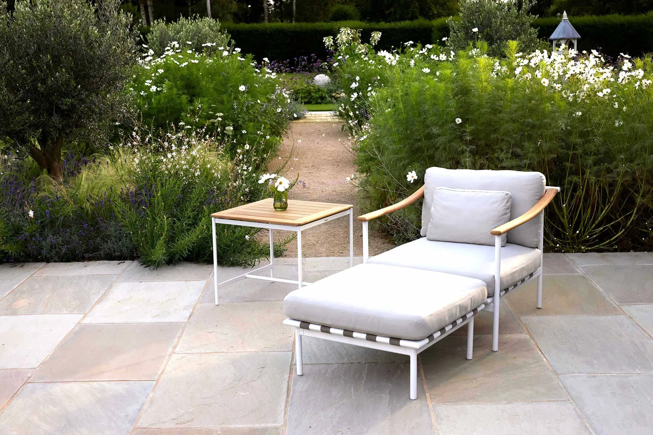 Outdoor patio with a modern lounge chair, cushion, small wooden side table with a vase containing a single white flower, surrounded by lush garden plants and flowers.