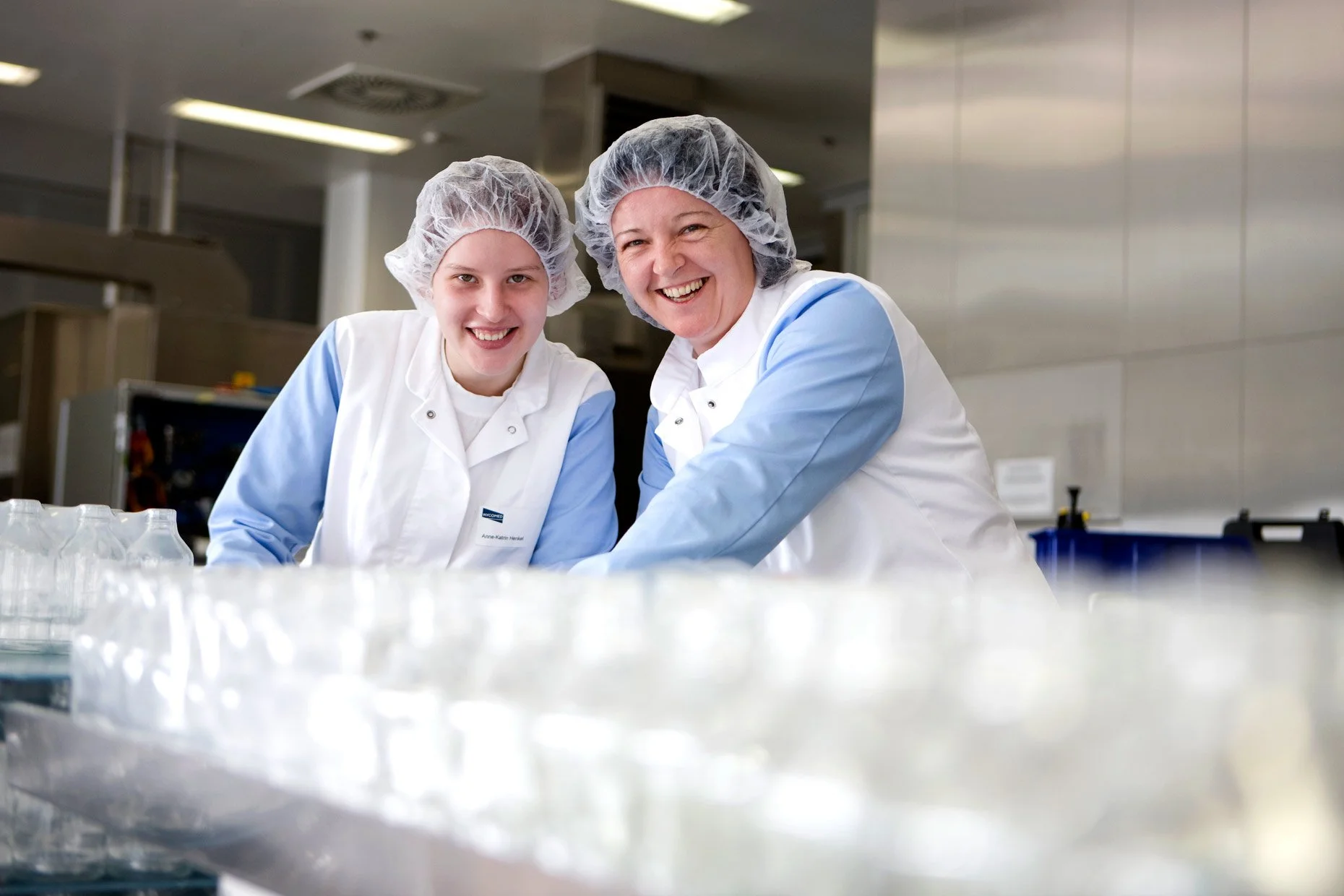 Two women wearing hairnets and white coats working in a laboratory or food processing facility, smiling at the camera.