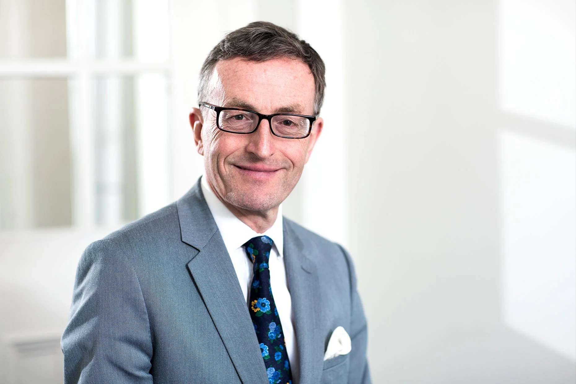 A middle-aged man wearing glasses in a gray suit, tie, and white shirt, smiling in an office setting.