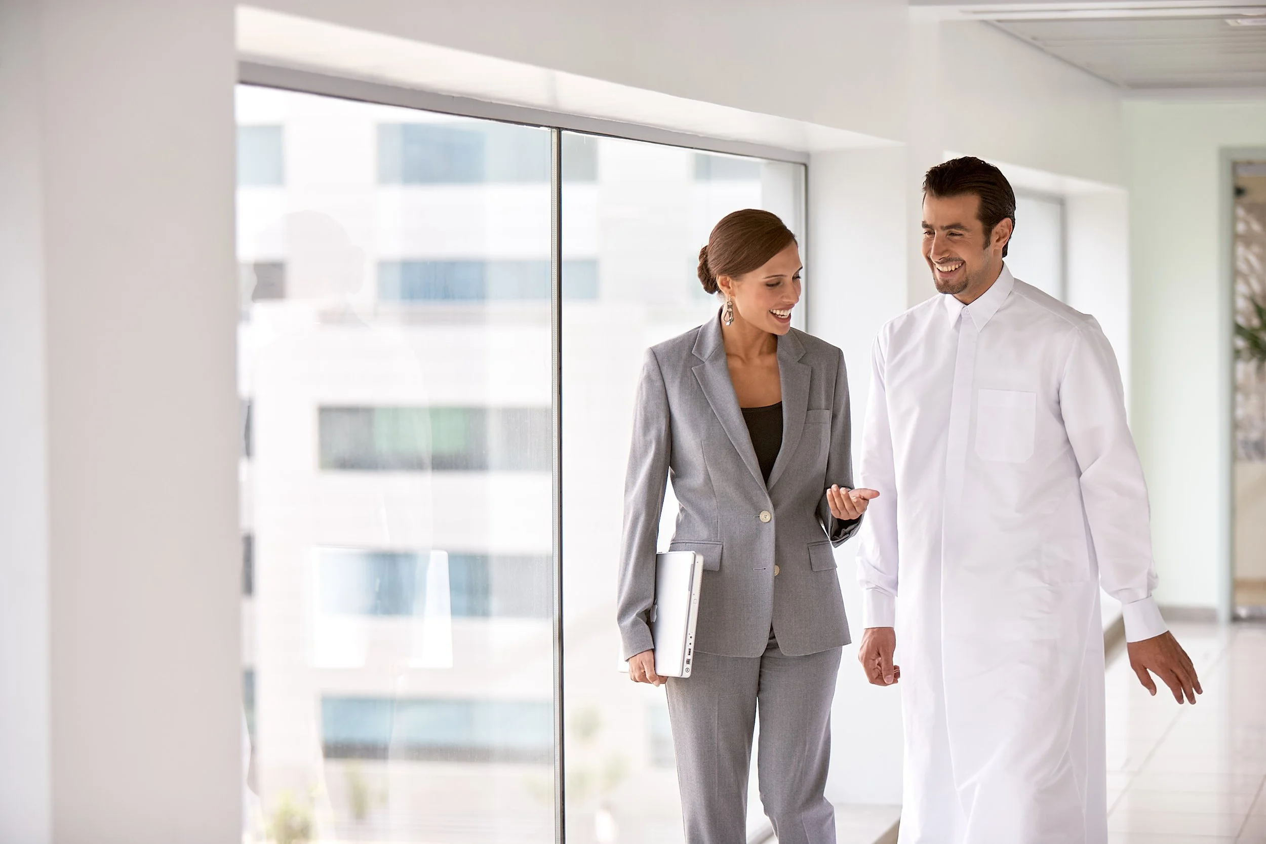 A man and a woman are walking together in an office building, smiling and engaged in conversation. The woman is dressed in a gray business suit and holding a laptop, while the man is wearing a white lab coat.