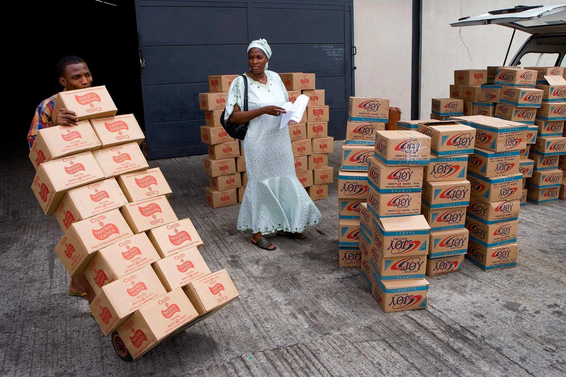 A market trader in Lagos standing outside as boxes are loaded for her business, reflecting entrepreneurship and daily trade in a bustling urban setting.