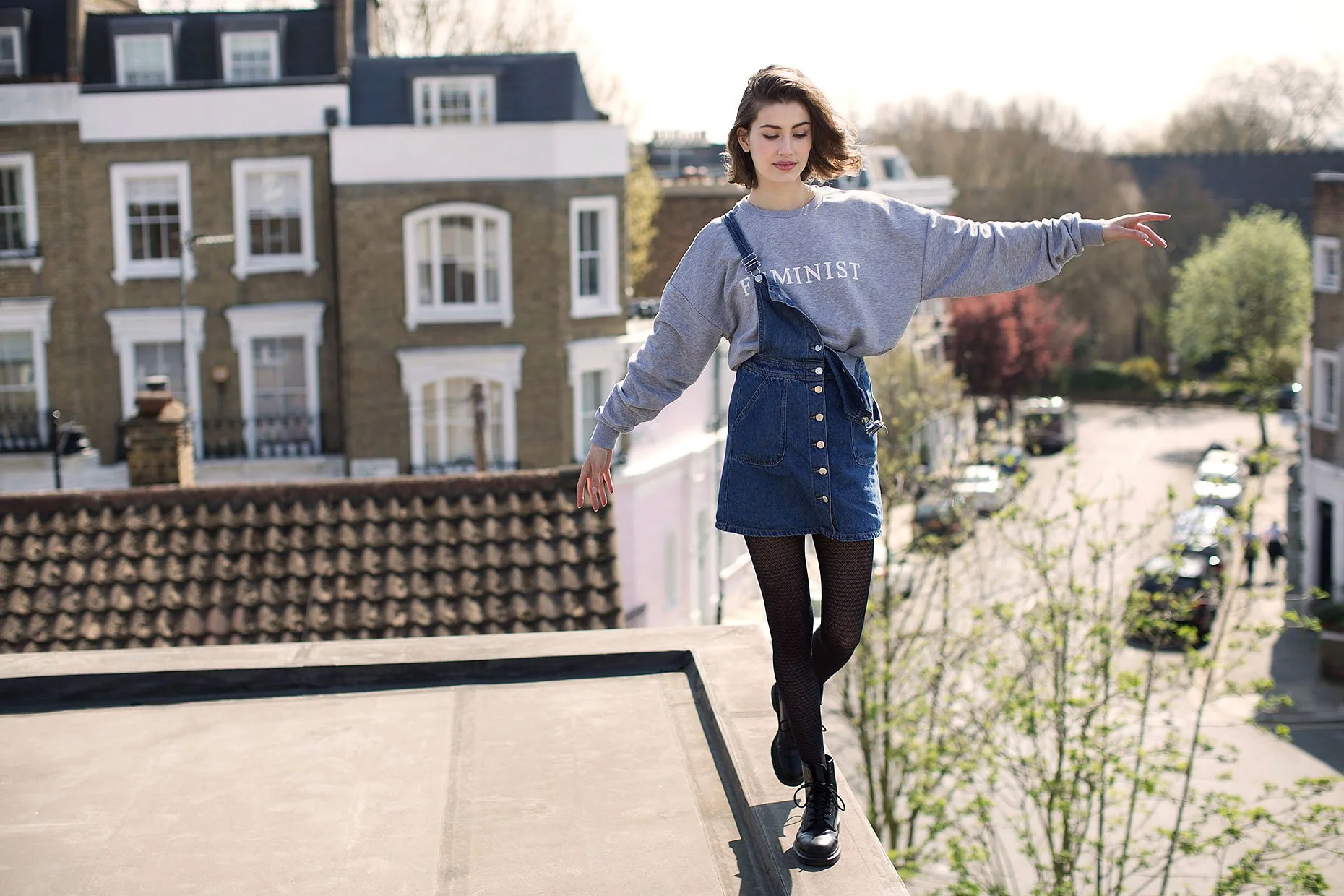 Person balancing on a ledge with arms outstretched, wearing a gray sweatshirt and denim dress. Background of residential buildings and trees.
