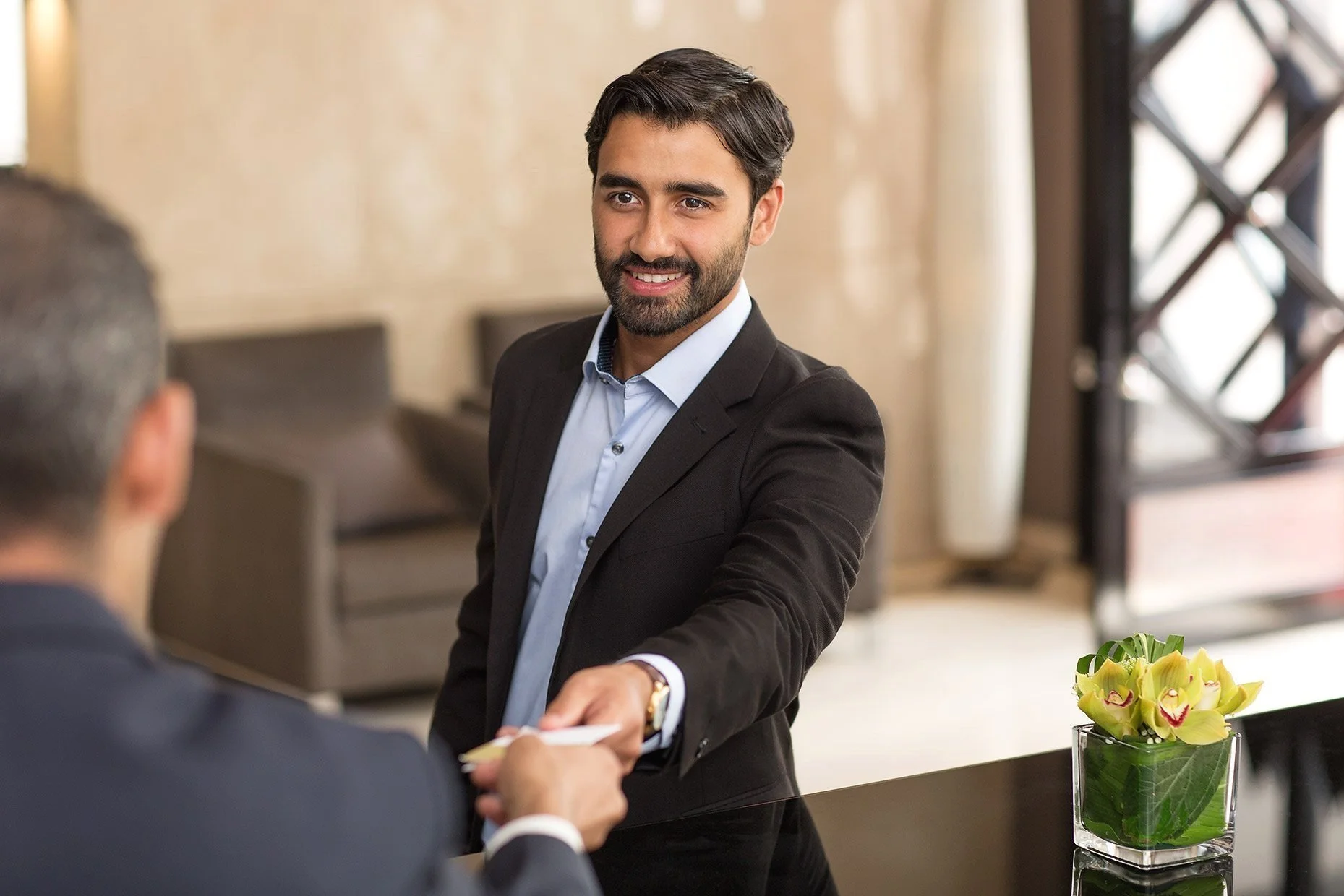 Man in business attire handing card to another person across a desk, with a vase of yellow flowers in the foreground.