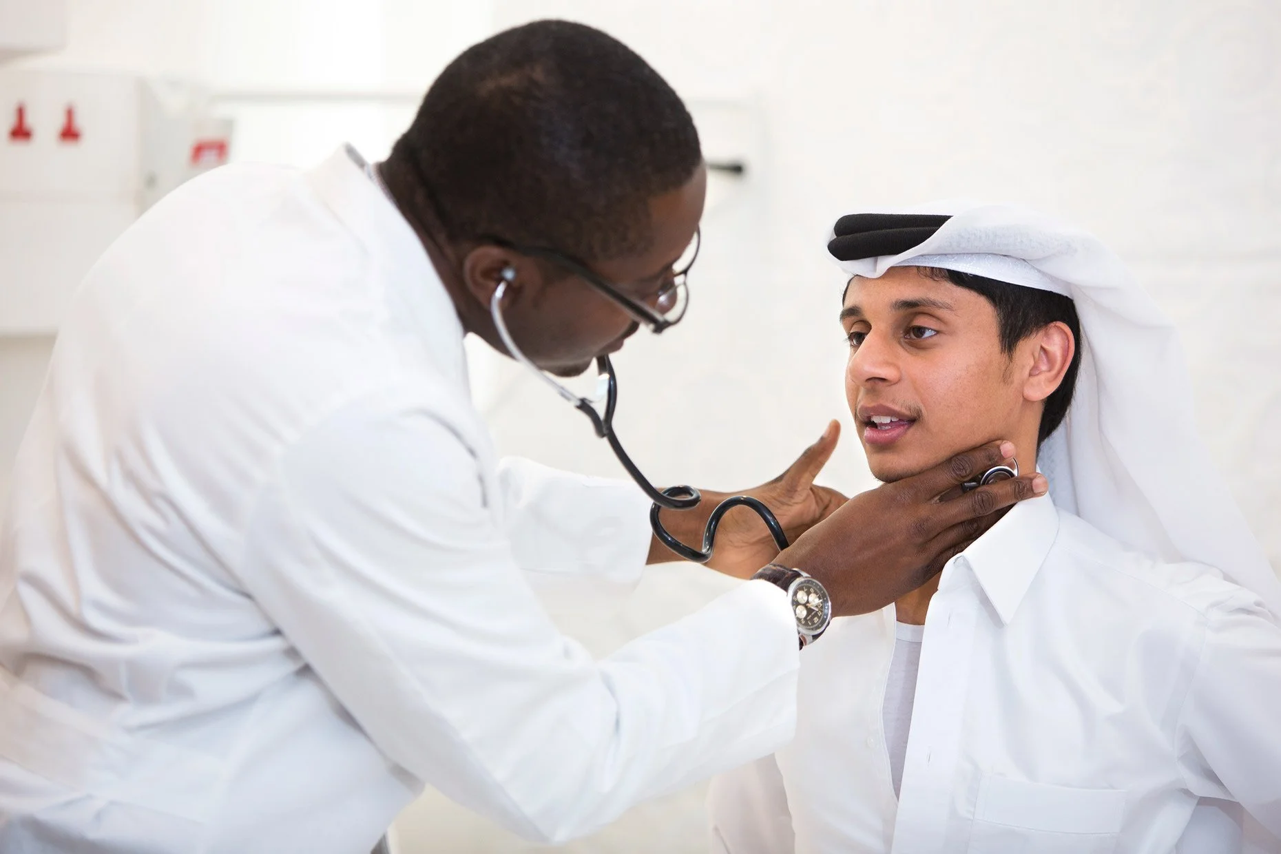 Doctor listening to young man's throat with a stethoscope in a medical setting.