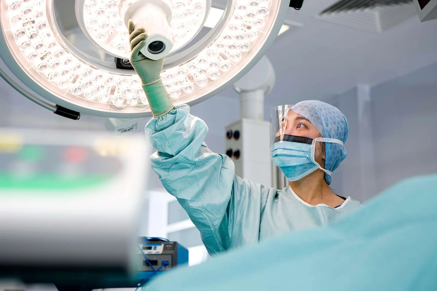 An ICU surgeon wearing a mask and adjusting an overhead surgical light, preparing for a procedure in a critical care setting.