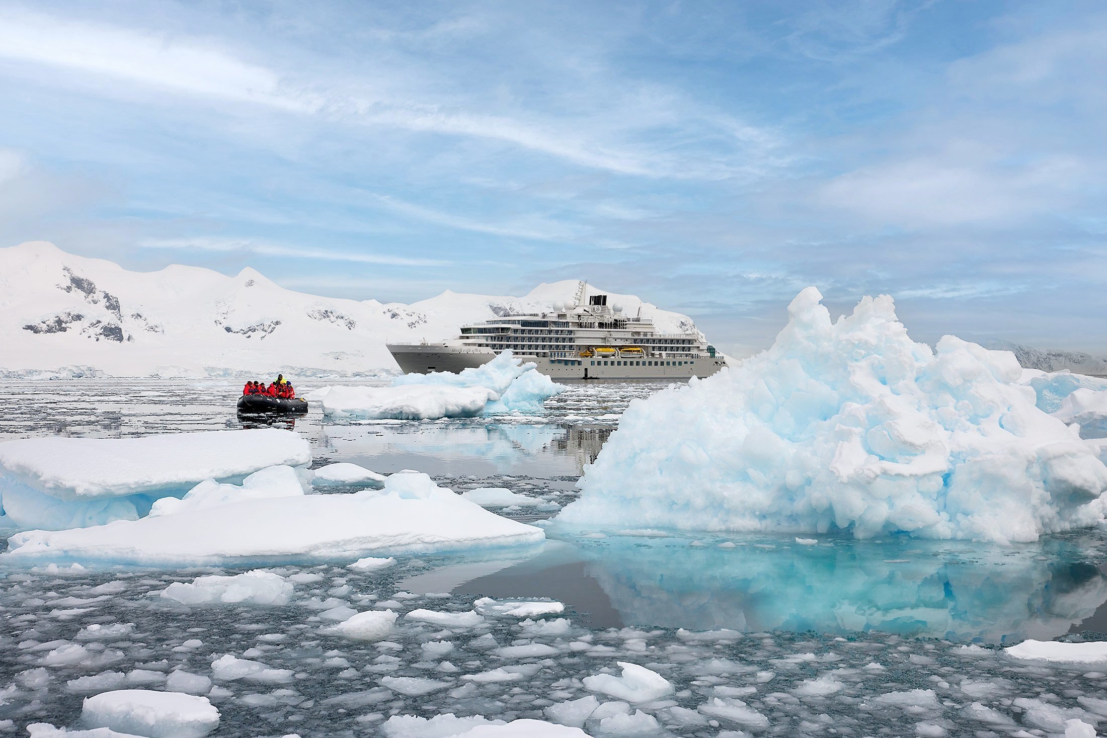 A cruise ship and a small boat with people in cold Antarctica waters, surrounded by icebergs and snowy landscape.