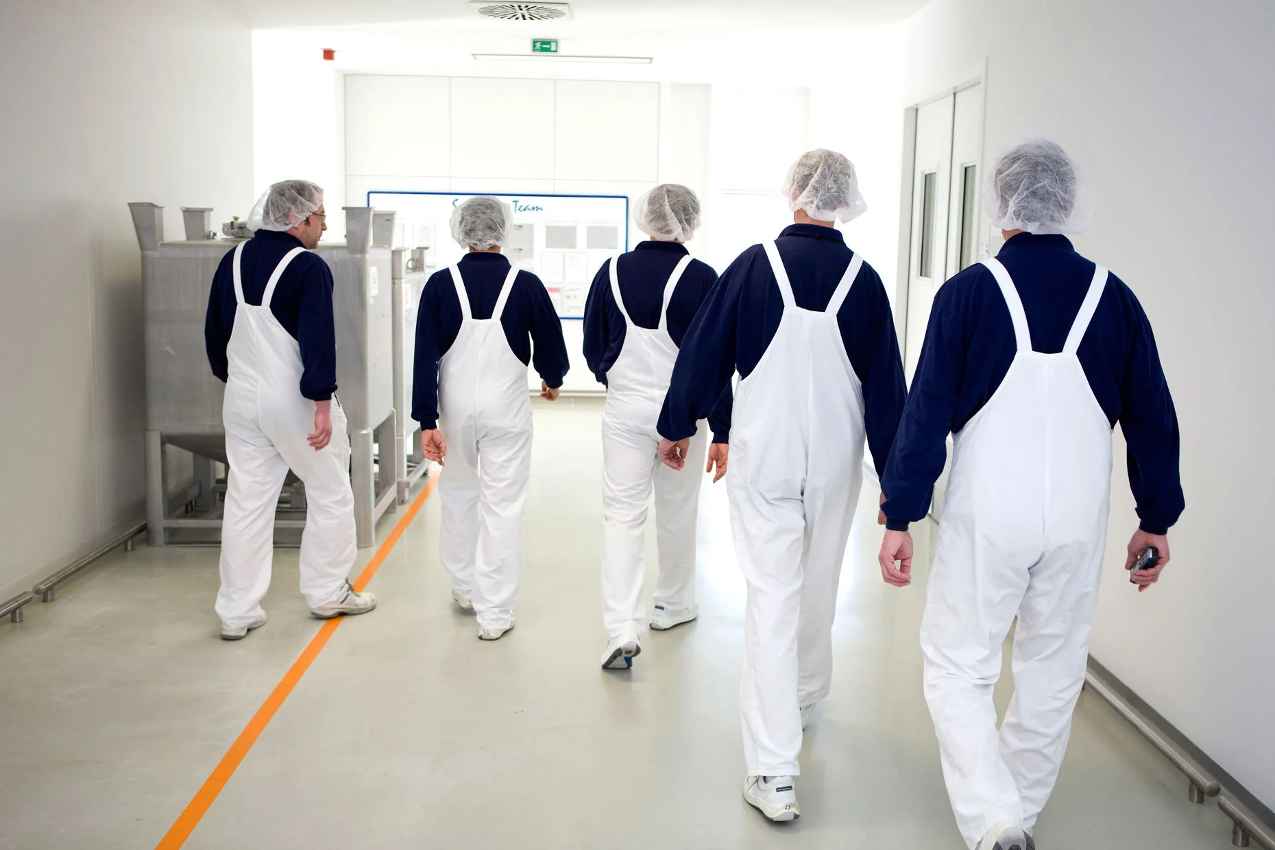 Group of people in lab coats, hair nets, and white aprons walking down a corridor in a laboratory or clean room