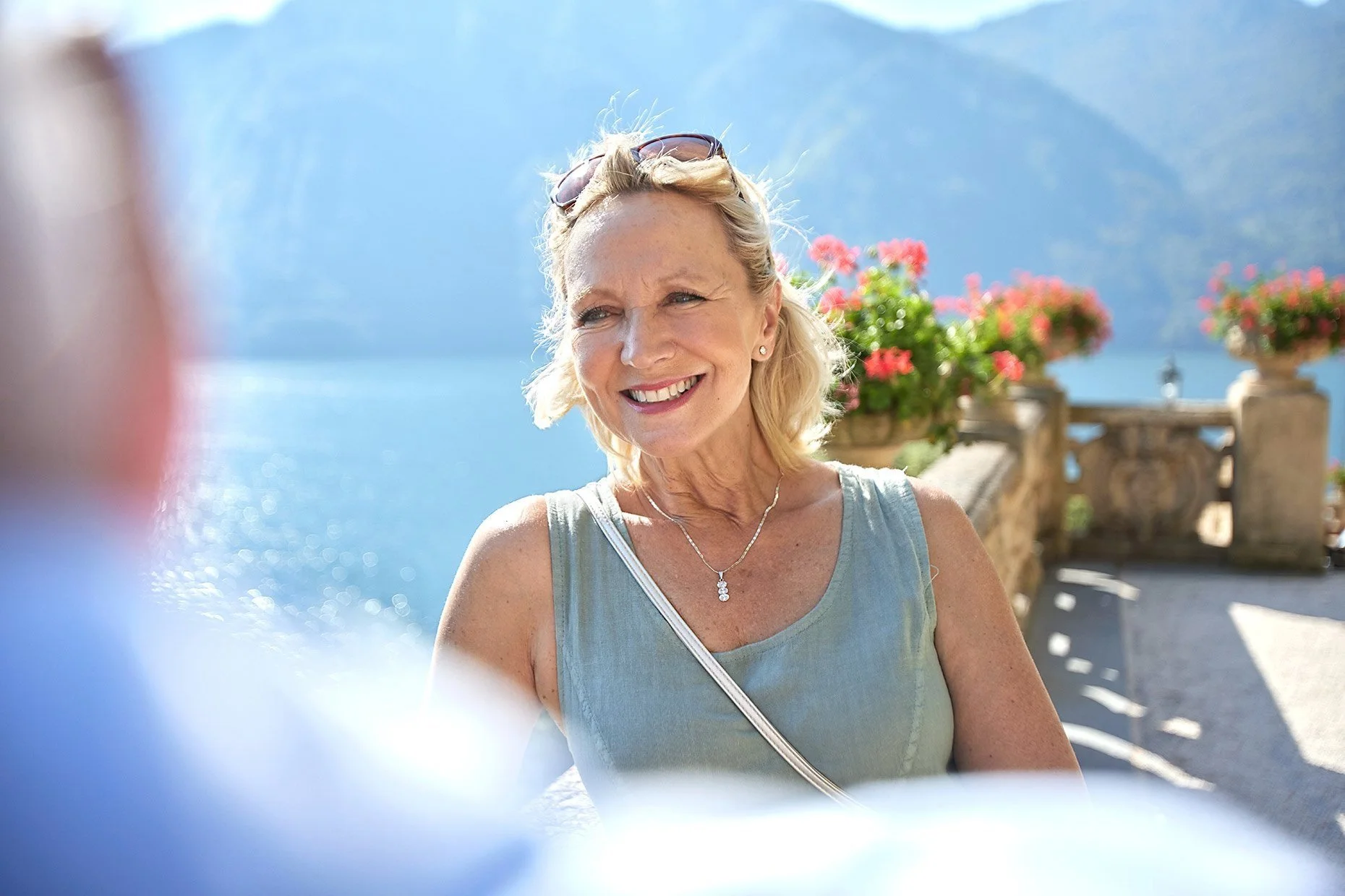 Smiling woman outdoors near water and mountains, wearing sunglasses on head, with flowers in background.