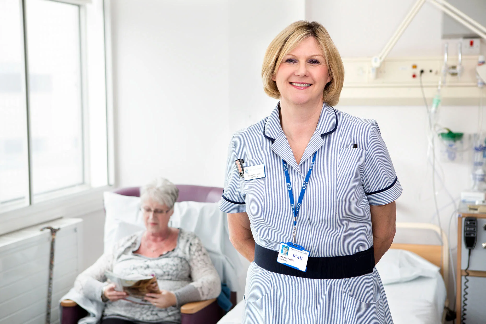 Head nurse looking confidently at the camera with a seated patient in the background, capturing professionalism and compassionate care in a healthcare setting.