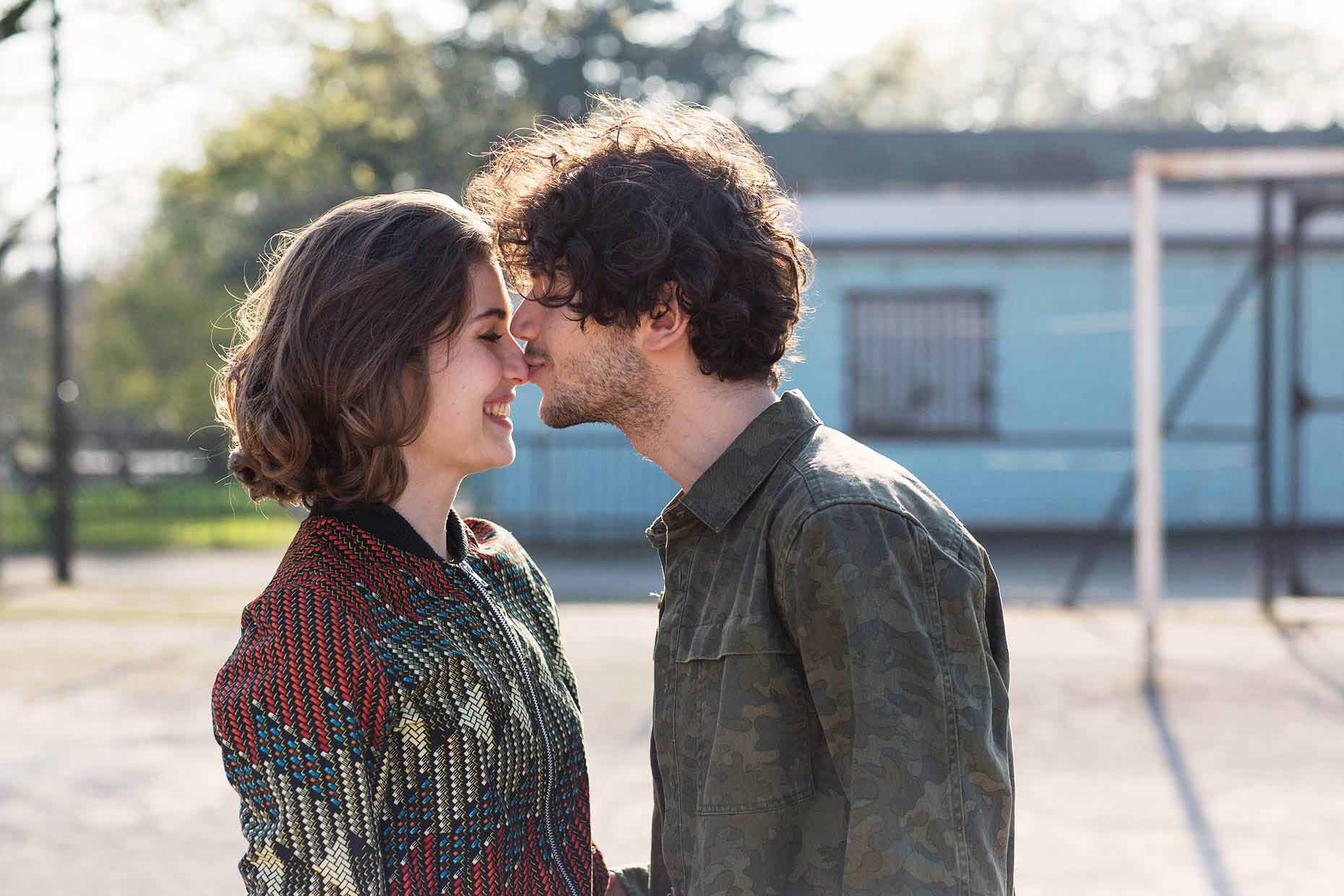 A couple in casual clothes touching noses outdoors, smiling at each other on a sunny day.