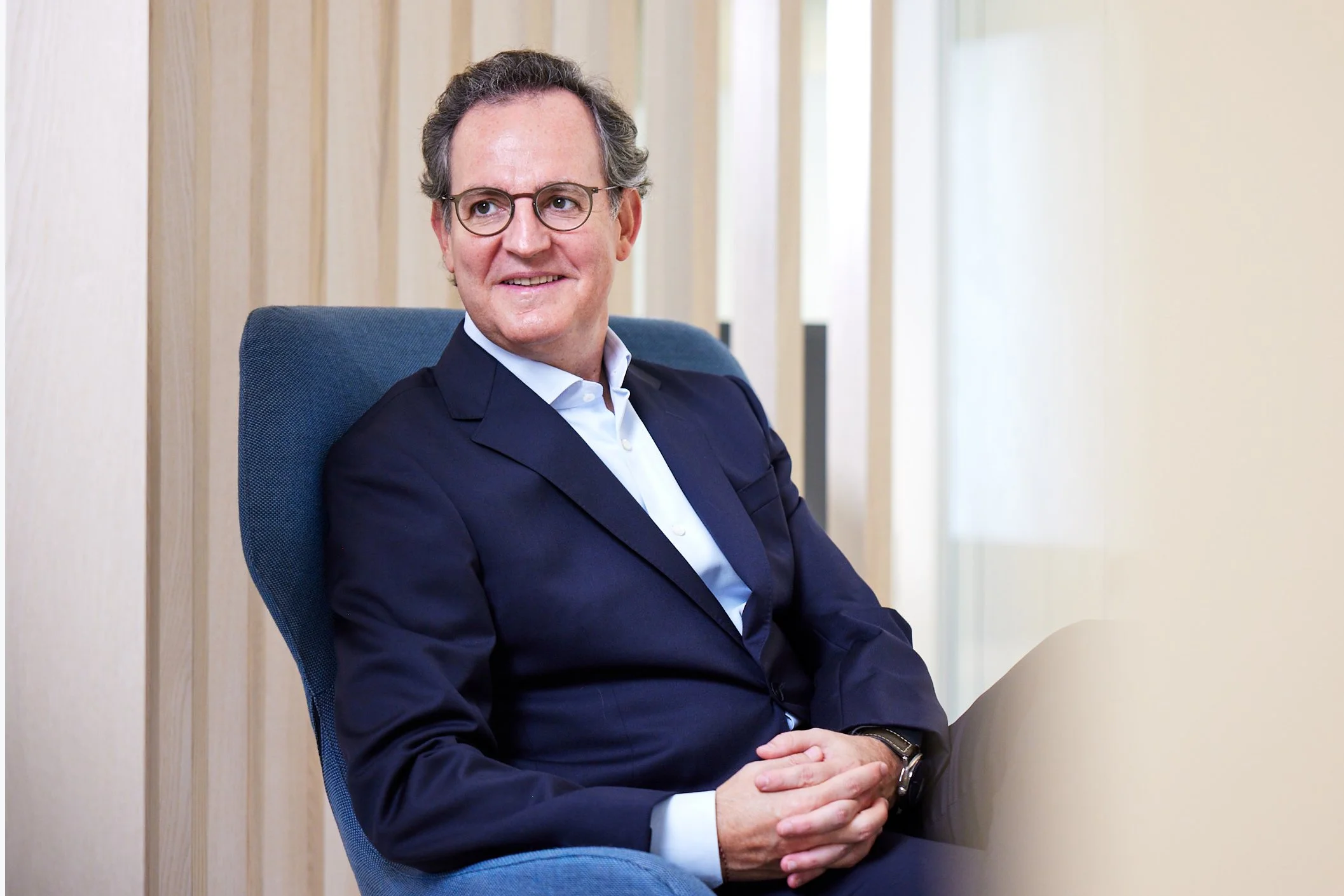 A man in a navy suit with a white shirt, sitting in an office chair with wooden wall panels behind him and natural light coming from the right.