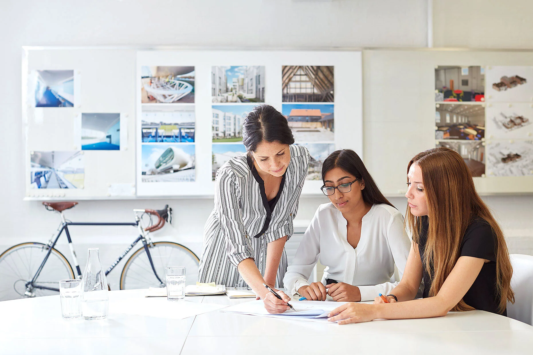 Three women collaborating in a modern office with architectural designs on the wall and a bicycle in the background.