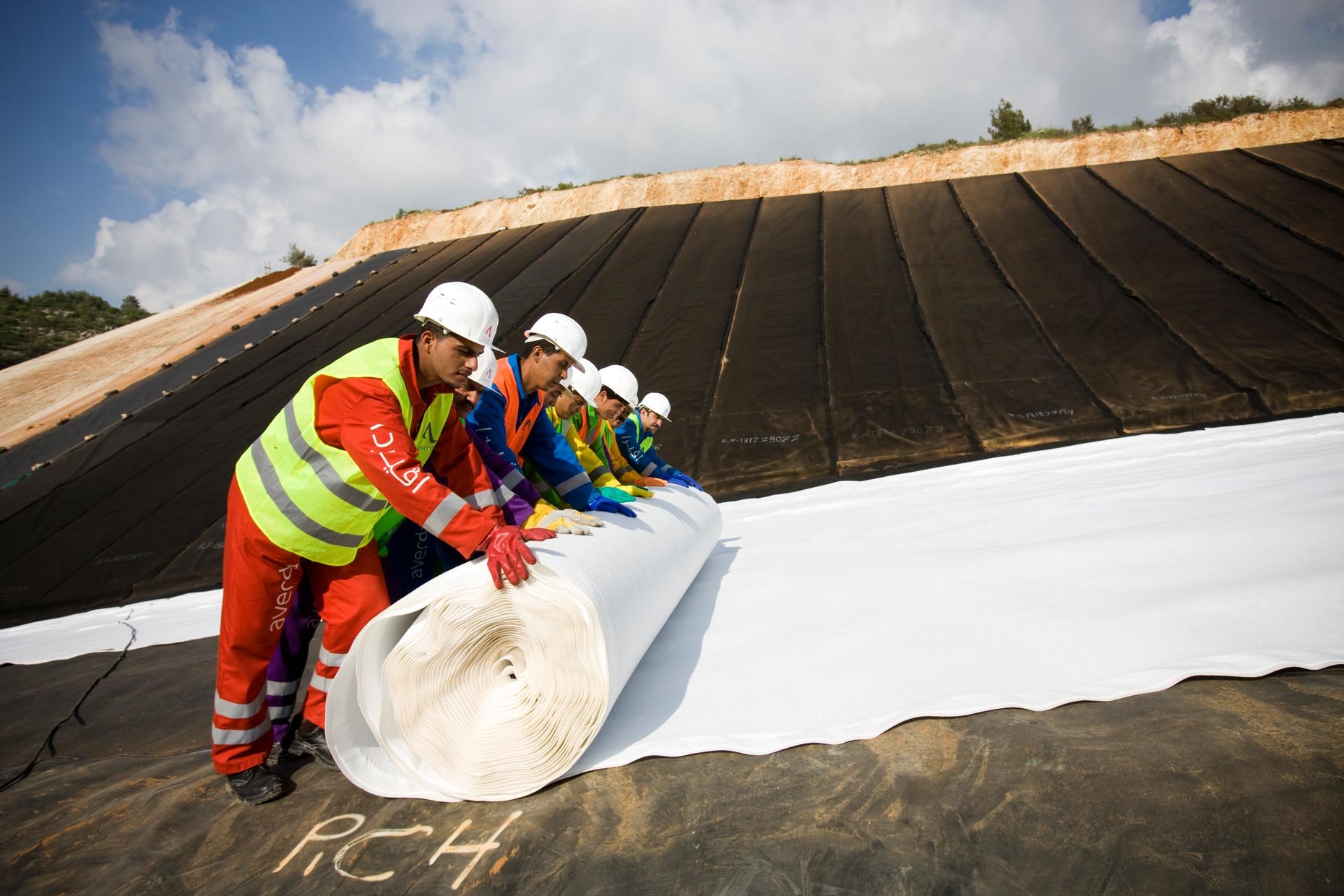 Group of construction workers in safety gear unrolling a large white waterproof membrane on a sloped roof under a cloudy sky.