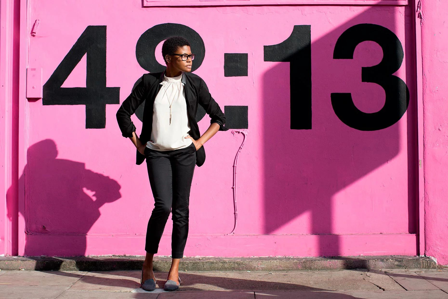 A stylish businesswoman dressed in black, standing confidently against a vibrant pink wall in a modern urban setting.